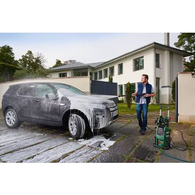Muž myje šedé auto vysokotlakým čističem před moderním bílým domem.