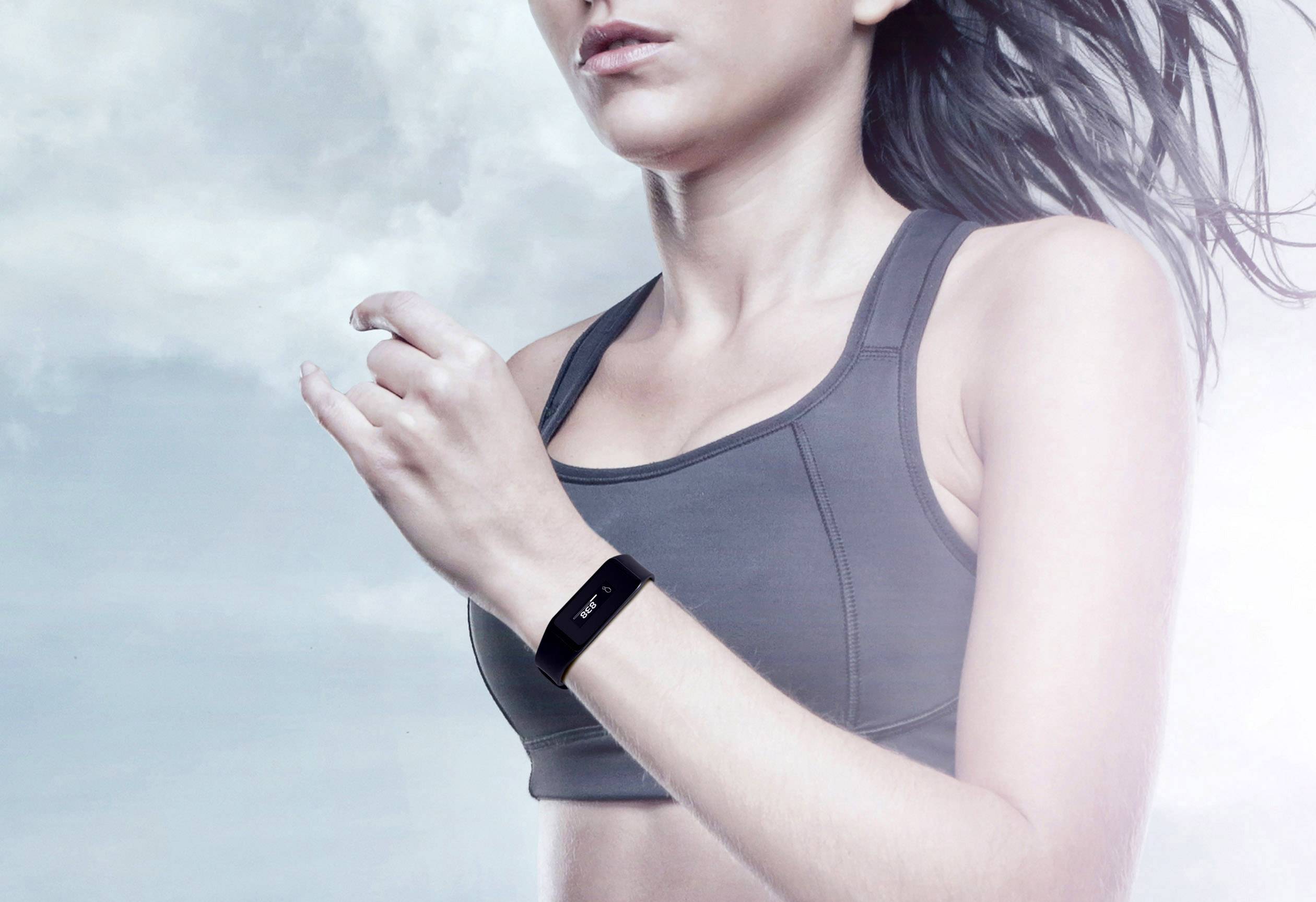 Žena ve sportovním oblečení nosí fitness náramek, zatímco cvičí venku. Na pozadí je zatažená obloha.