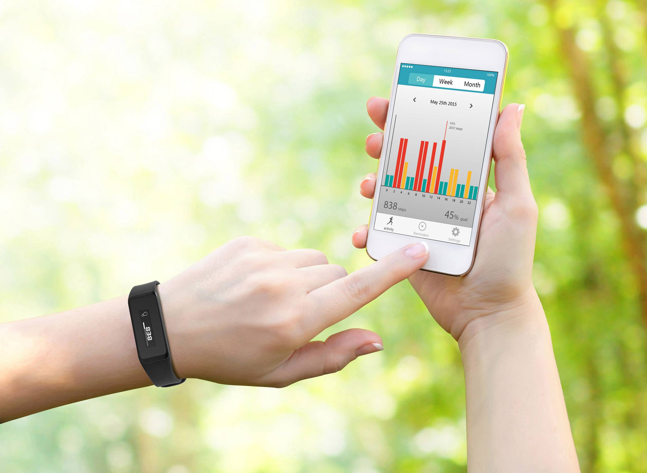 Jedna ruka drží smartphone s fitness aplikací, která zobrazuje sloupcové grafy. Druhá ruka má nasazený fitness náramek, obklopena lesem.