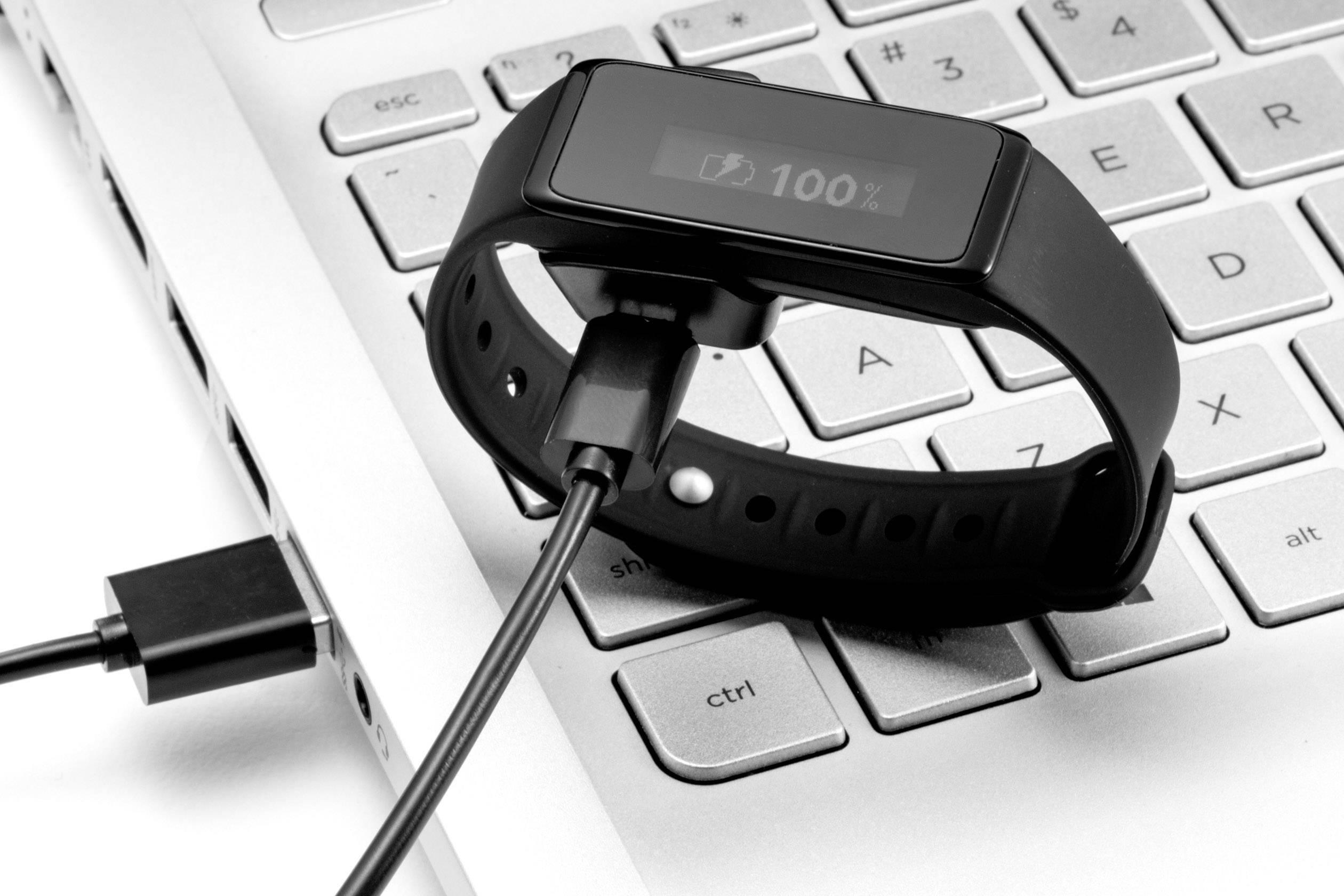 Fitness tracker je nabíjen přes USB kabel na klávesnici notebooku. Display ukazuje '100 %', což signalizuje úplné nabití.