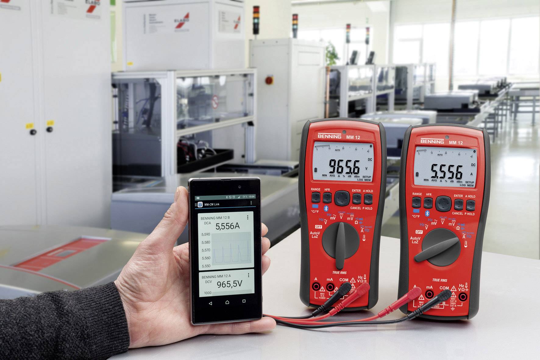 Jedna ruka drží smartphone s čísly, vedle něj stojí dva digitální multimetry na stole v laboratorním prostředí.