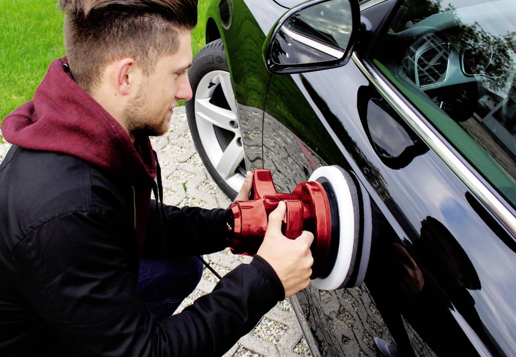 Muž leští auto červeným leštícím strojem, zatímco stojí na chodníku vedle černého automobilu.