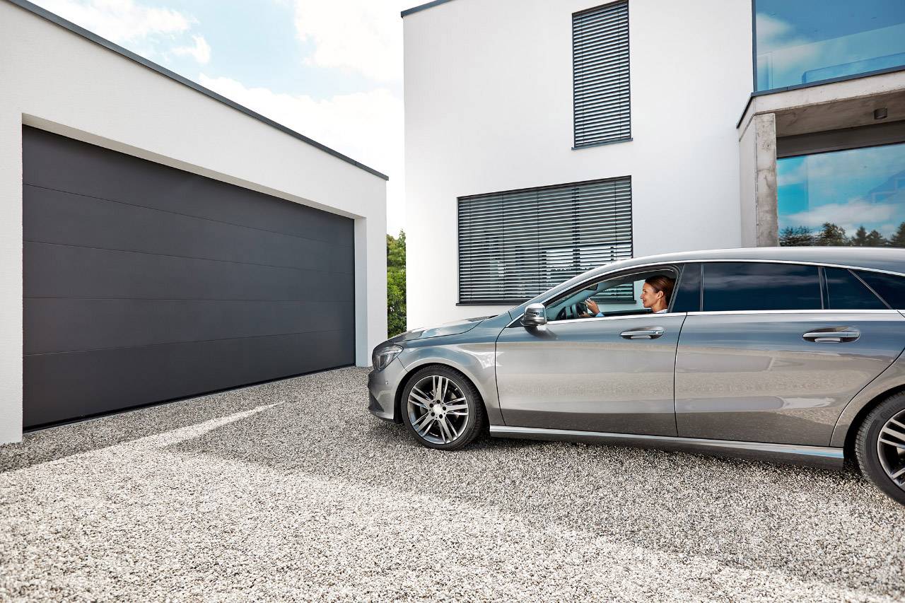 Stříbrné auto před moderním domem a garáží na štěrkové ploše. Řidič se dívá z okna směrem ke garáži.