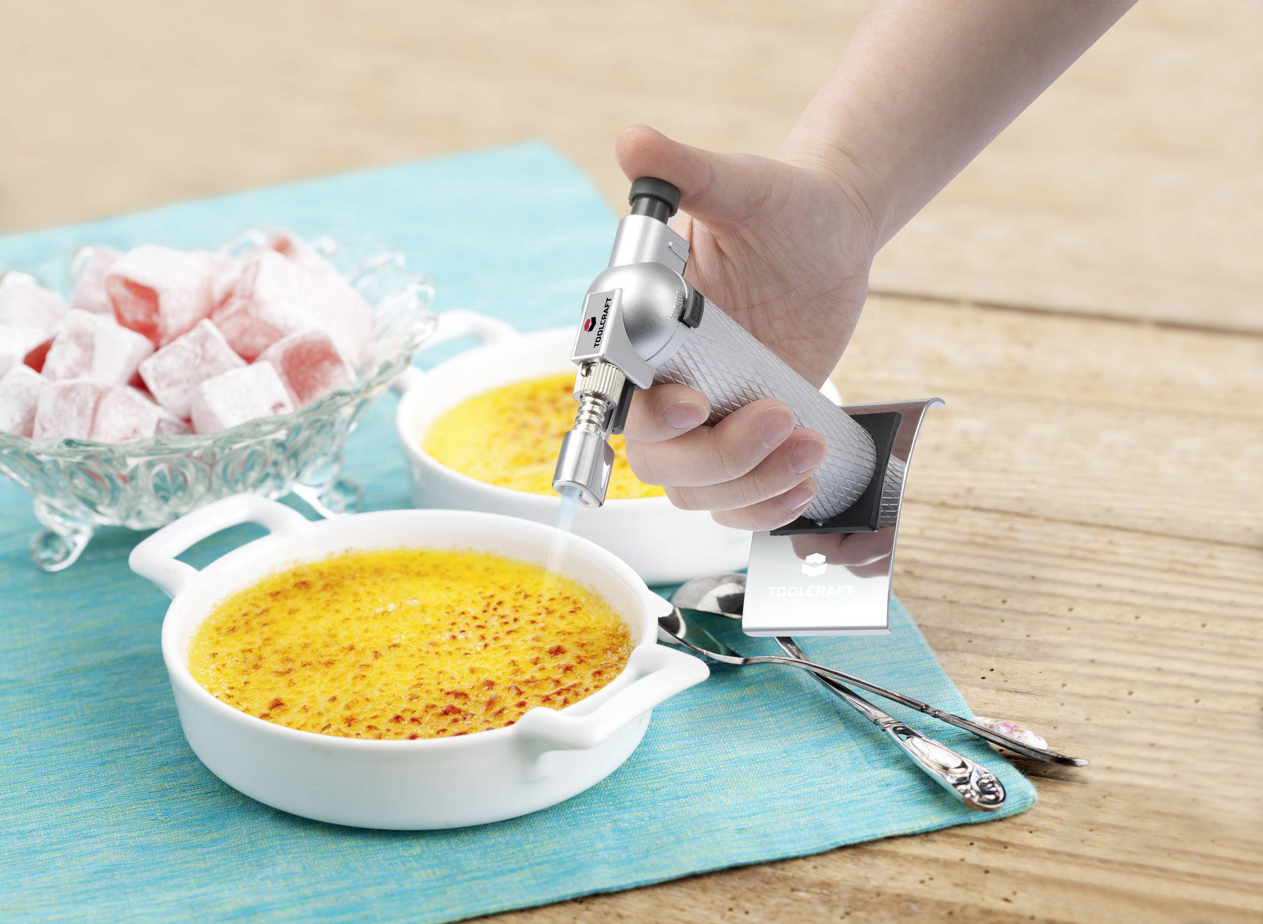 Ruka zapaluje horní vrstvu dvou crème brûlée pomocí flambovacího hořáku. Vedle stojí miska s tureckým potěšením (Turkish delights).