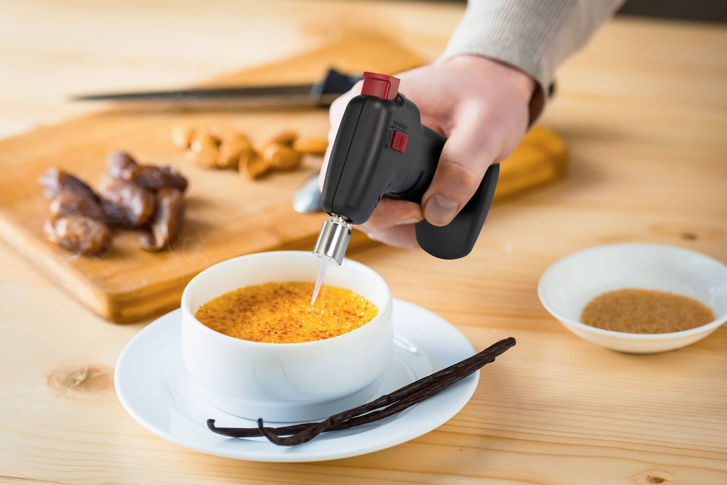 Osoba karamelizuje povrch crème brûlée malým plynovým hořákem. V pozadí leží koření a datle.