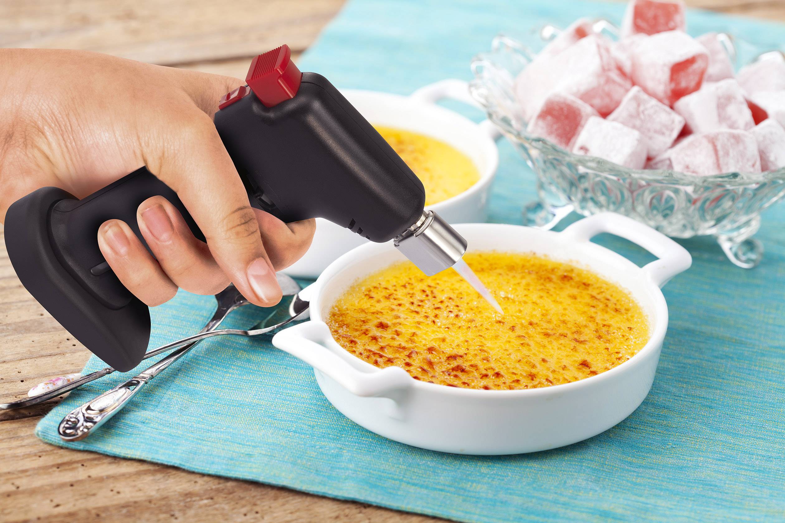 Ruka drží hořák na karamelizaci nad miskou s crème brûlée, vedle je miska s kostkami posypanými moučkovým cukrem.