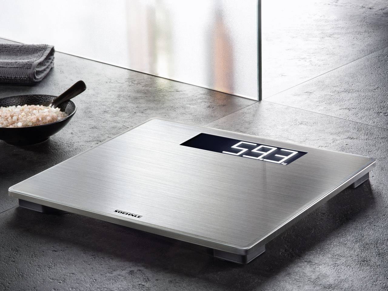 Digitální váha na šedé podlaze ukazuje hmotnost 59,3 kg. Vedle ní stojí miska s růžovou solí a ručník.