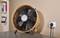 Stolní ventilátor v retro designu stojí na šedém stole. Je zapojen do elektrické sítě a připraven k použití. V pozadí je knihovna.