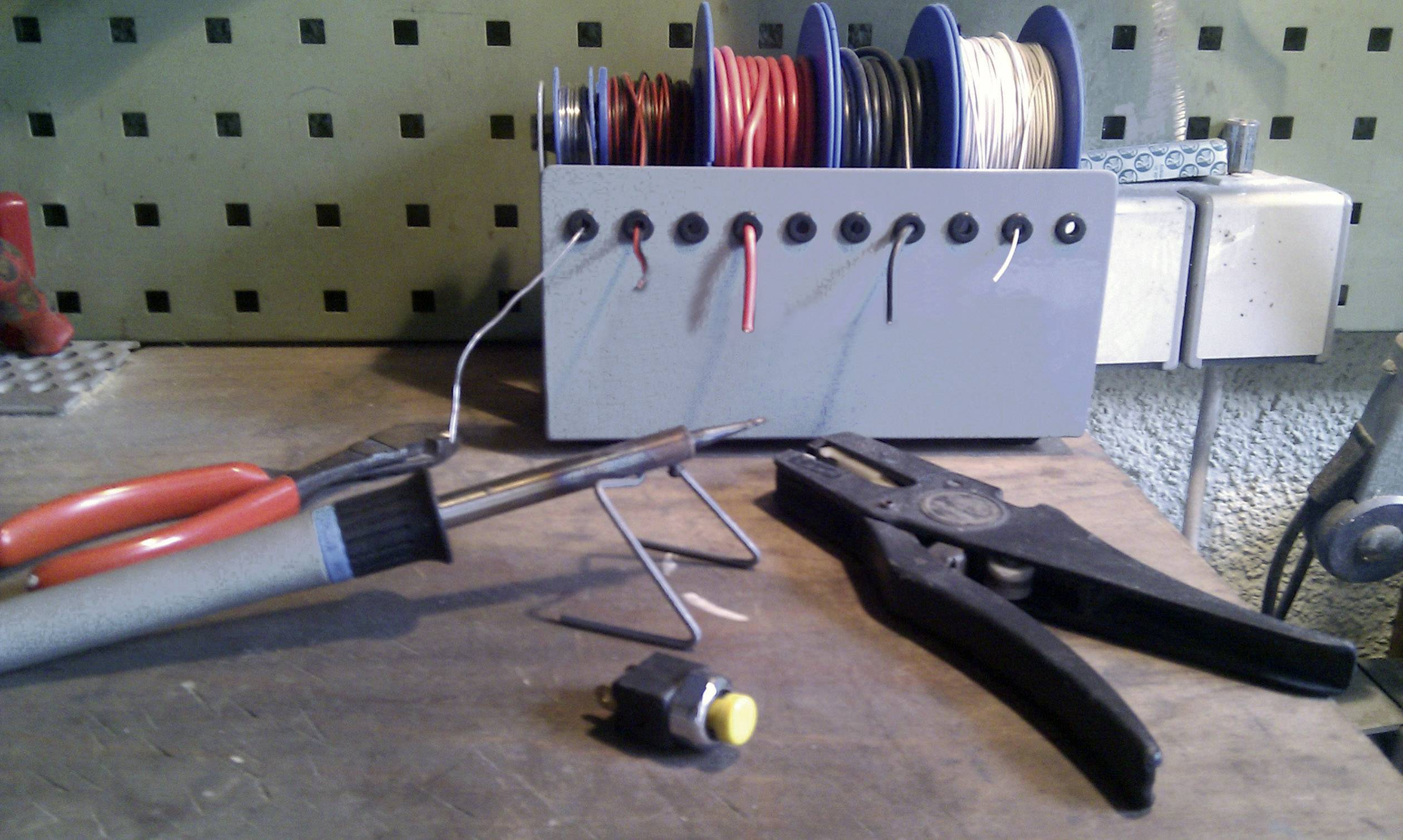 Pracovní stůl s pájecí pistolí, kleštěmi a cívkami drátu. Přepínač leží na dřevěném povrchu. Pozadí s děrovanou deskou.