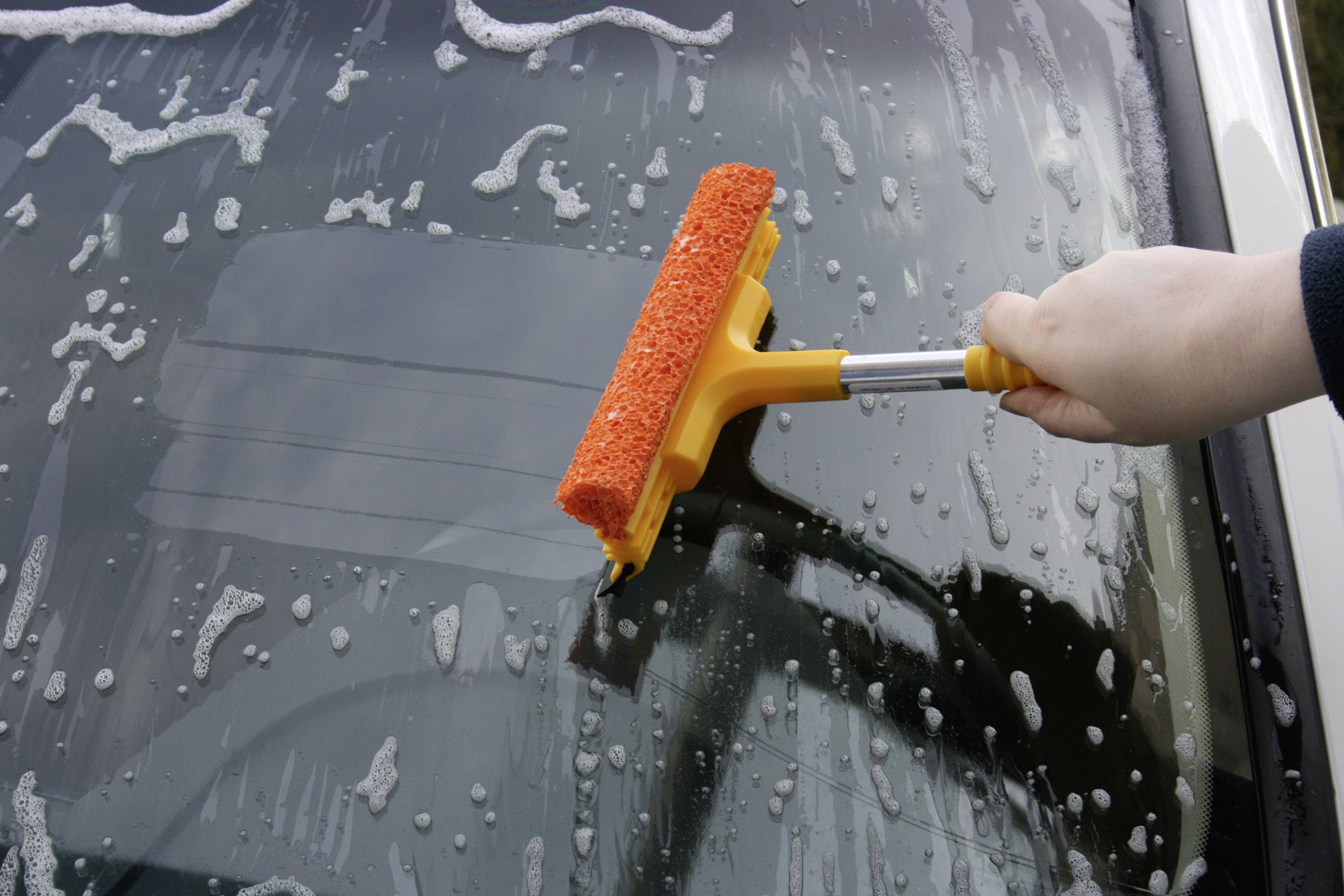 Osoba čistí čelní sklo automobilu žlutou stěrkou, přičemž je rozstříkána mýdlová voda.