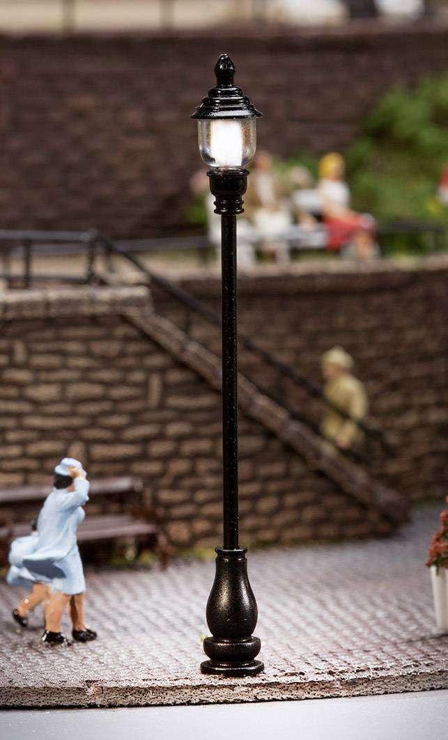 Pouliční lampa v Miniaturním městě, osvětlená. V pozadí rozmazané postavy na schodech a lavičce a zelené plochy.