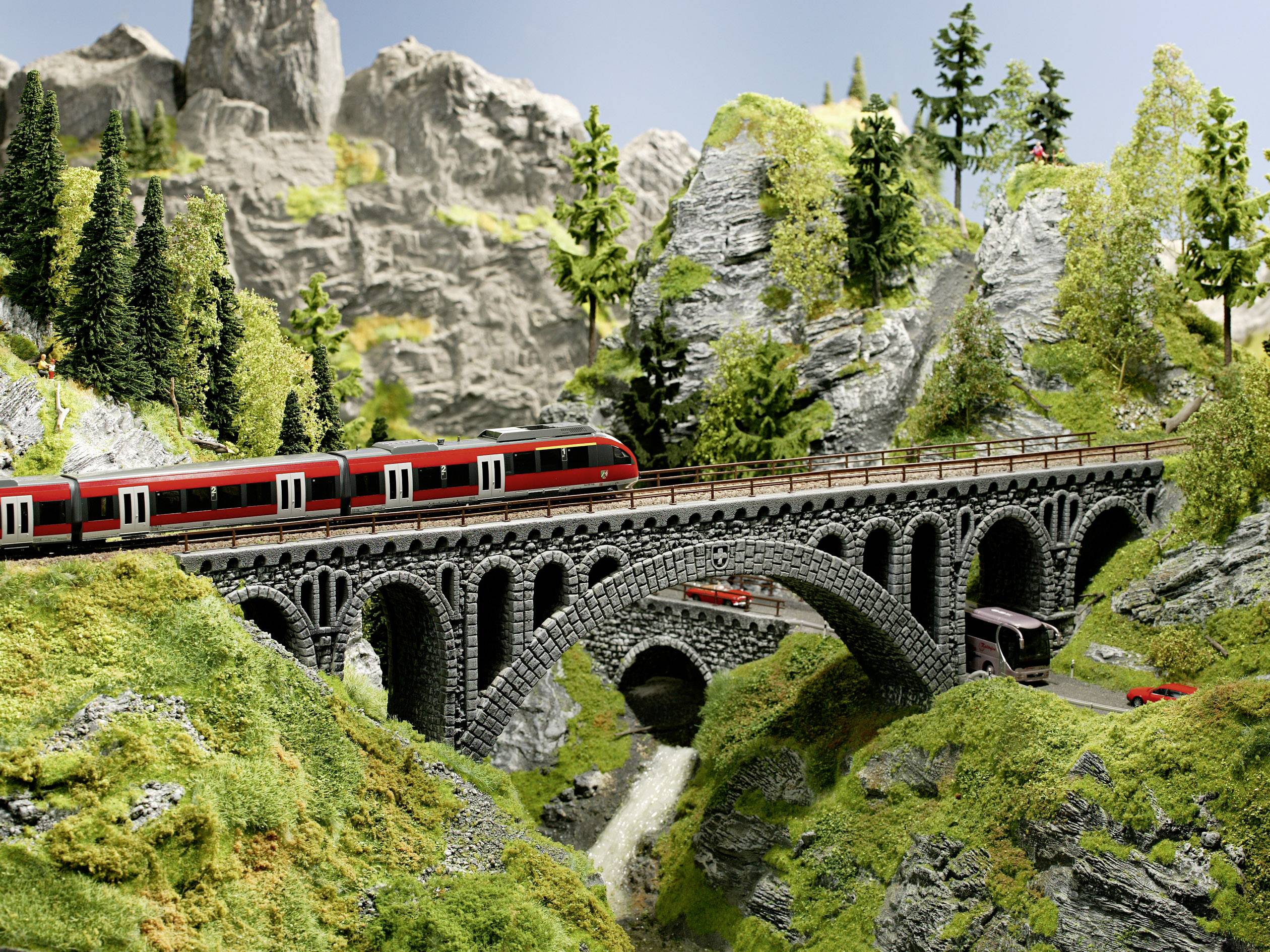 Červený vlak jede přes kamenný most v detailně propracované modelové krajině s horami, stromy a vodopádem pod ním.