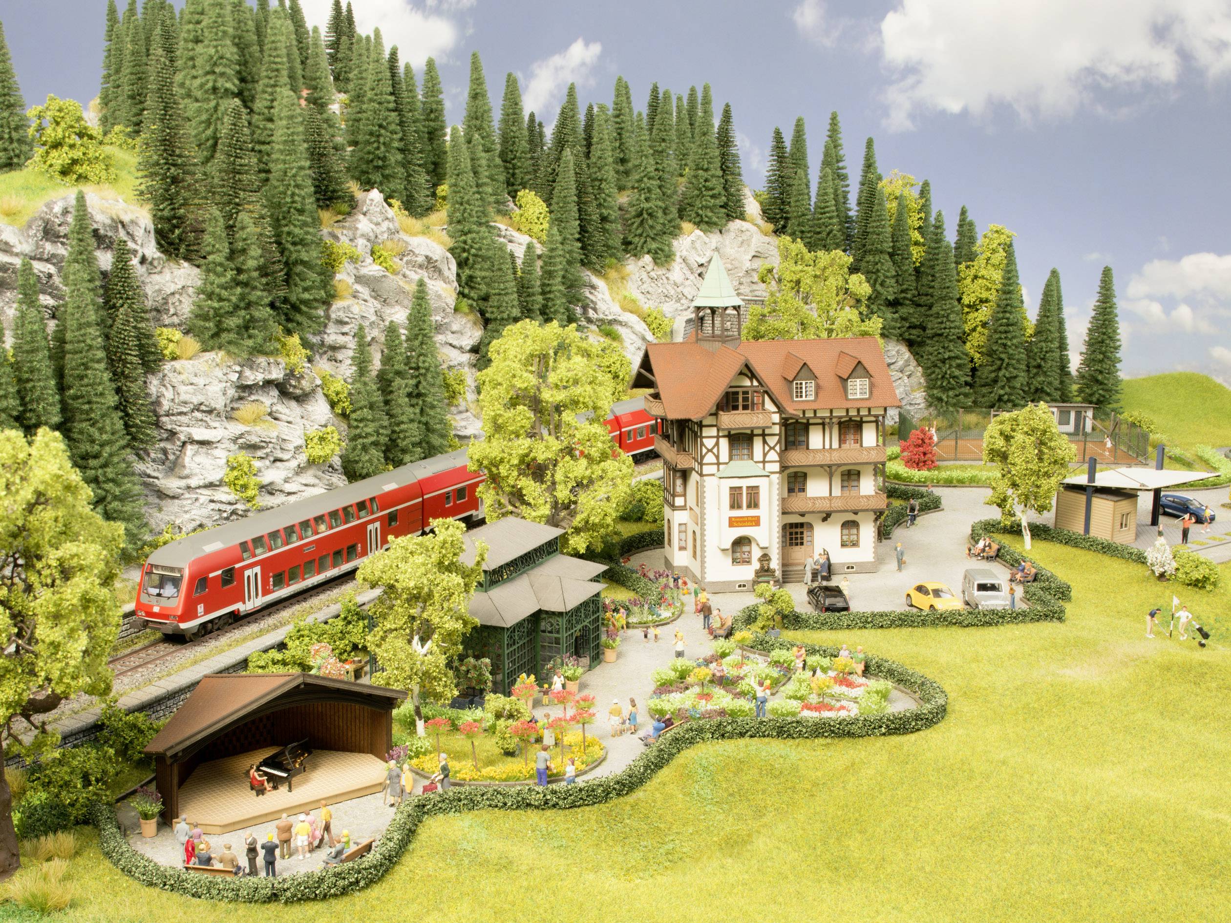 Miniaturní krajina s červeným modelovým vlakem projíždí kolem budovy ve stylu alpské architektury, obklopená stromy a lidmi.