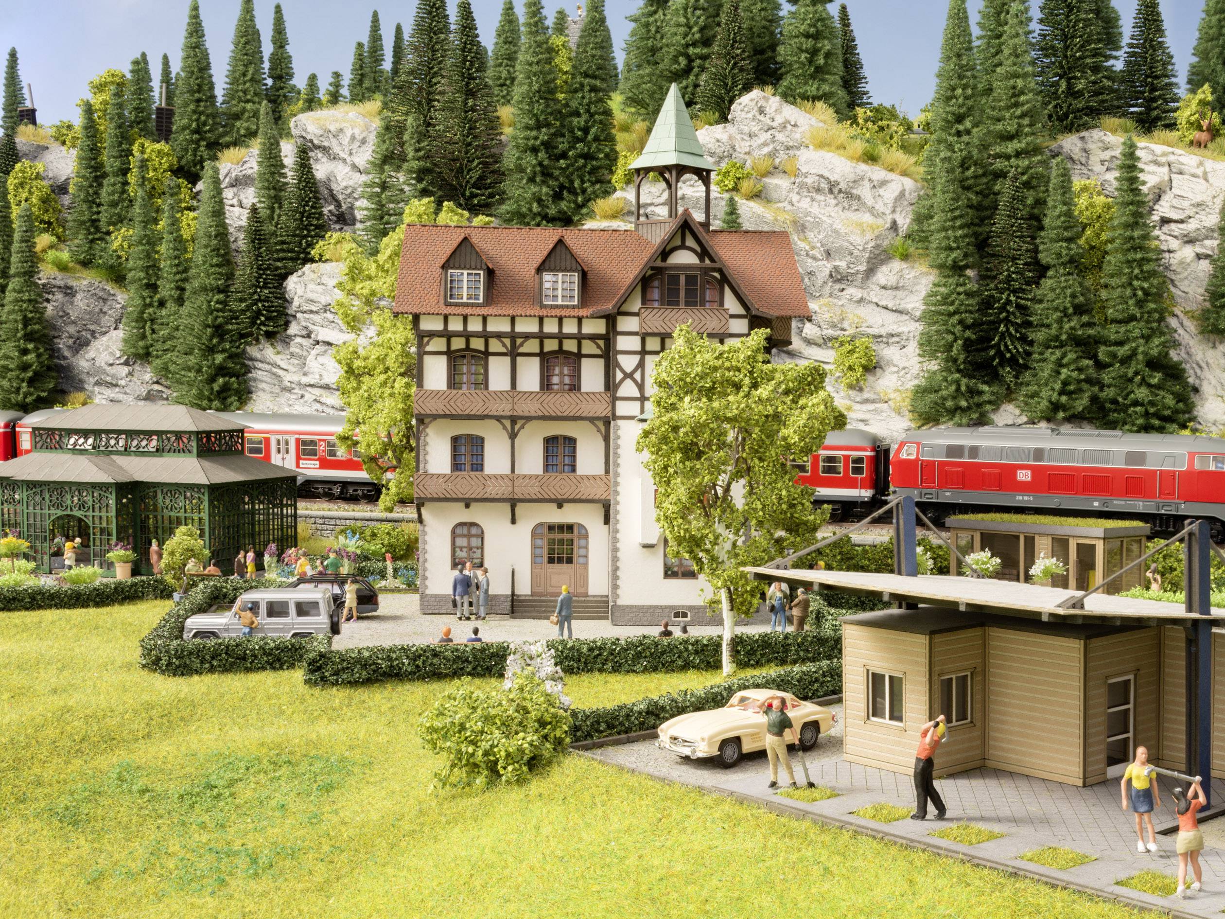 Miniaturní model zobrazuje nádraží v venkovské krajině s projíždějícím červeným vlakem, obklopeným stromy a kopci.