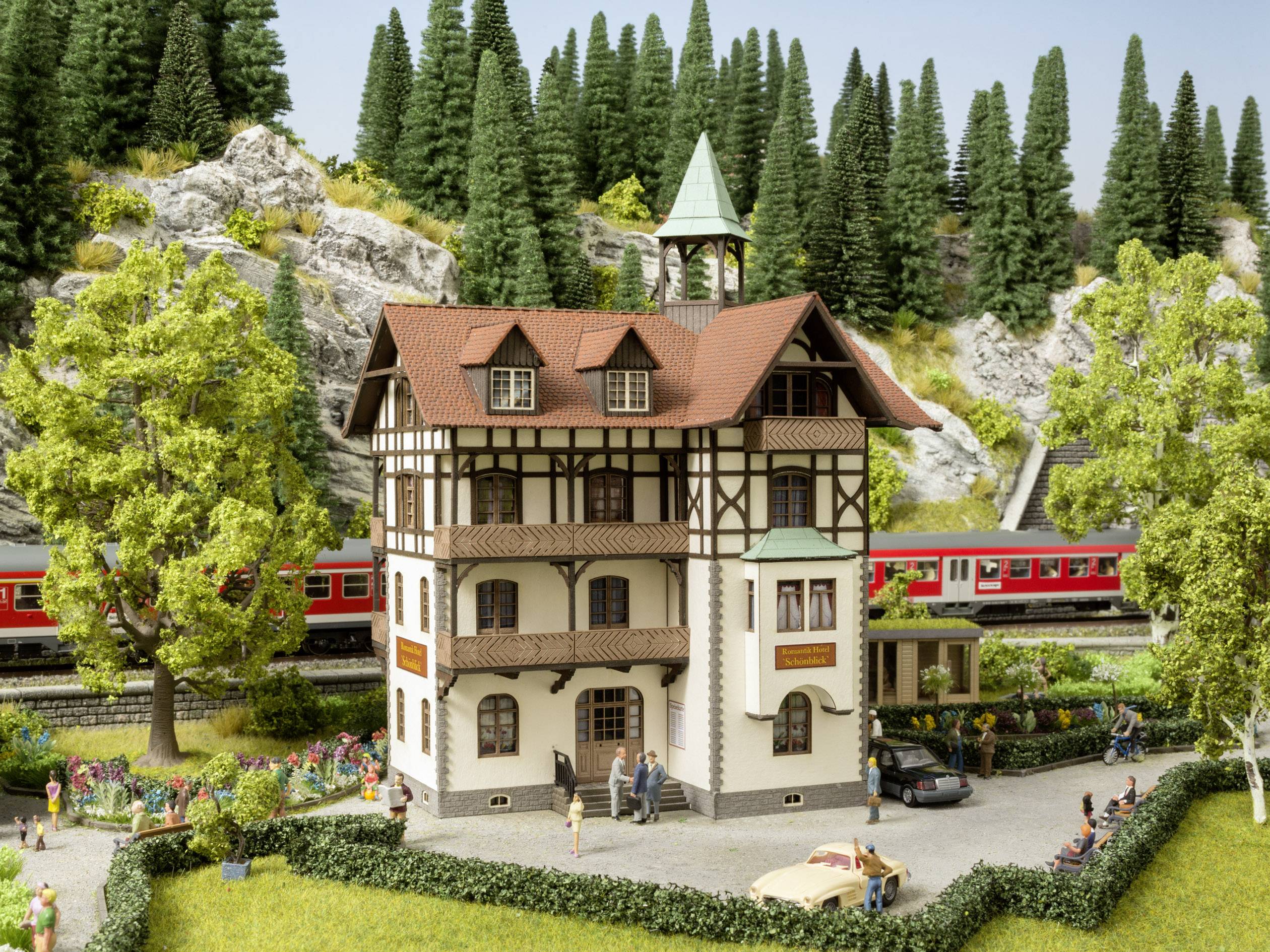 Miniaturní model zobrazuje hrázděnou budovu s věží, obklopenou stromy. V pozadí projíždí červený vlak.