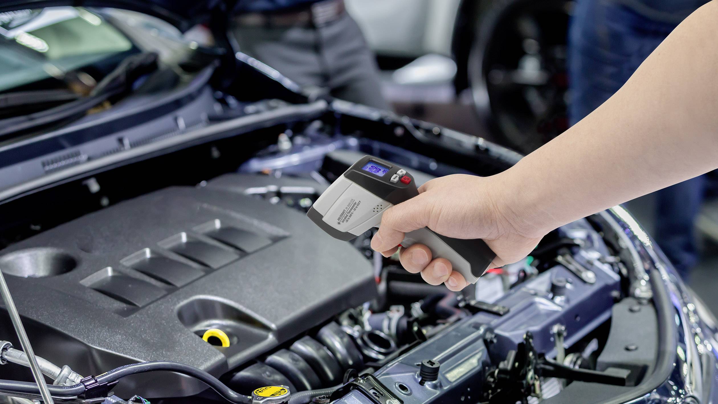 Jedna ruka drží digitální teploměr nad otevřeným motorovým prostorem automobilu za účelem měření teploty.