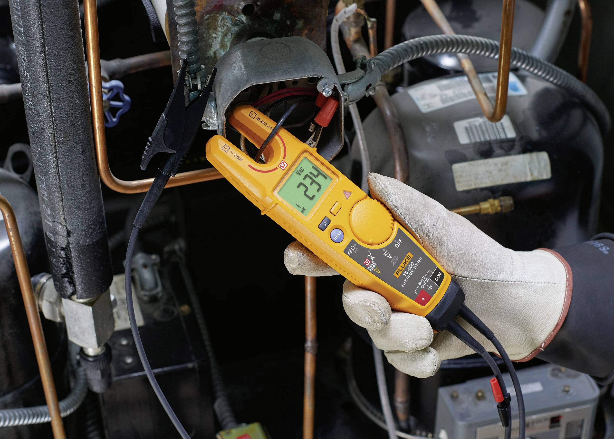 Osoba drží digitální multimetr, který ukazuje 234 voltů na elektrické instalaci. V pozadí jsou měděné trubky a kabely.