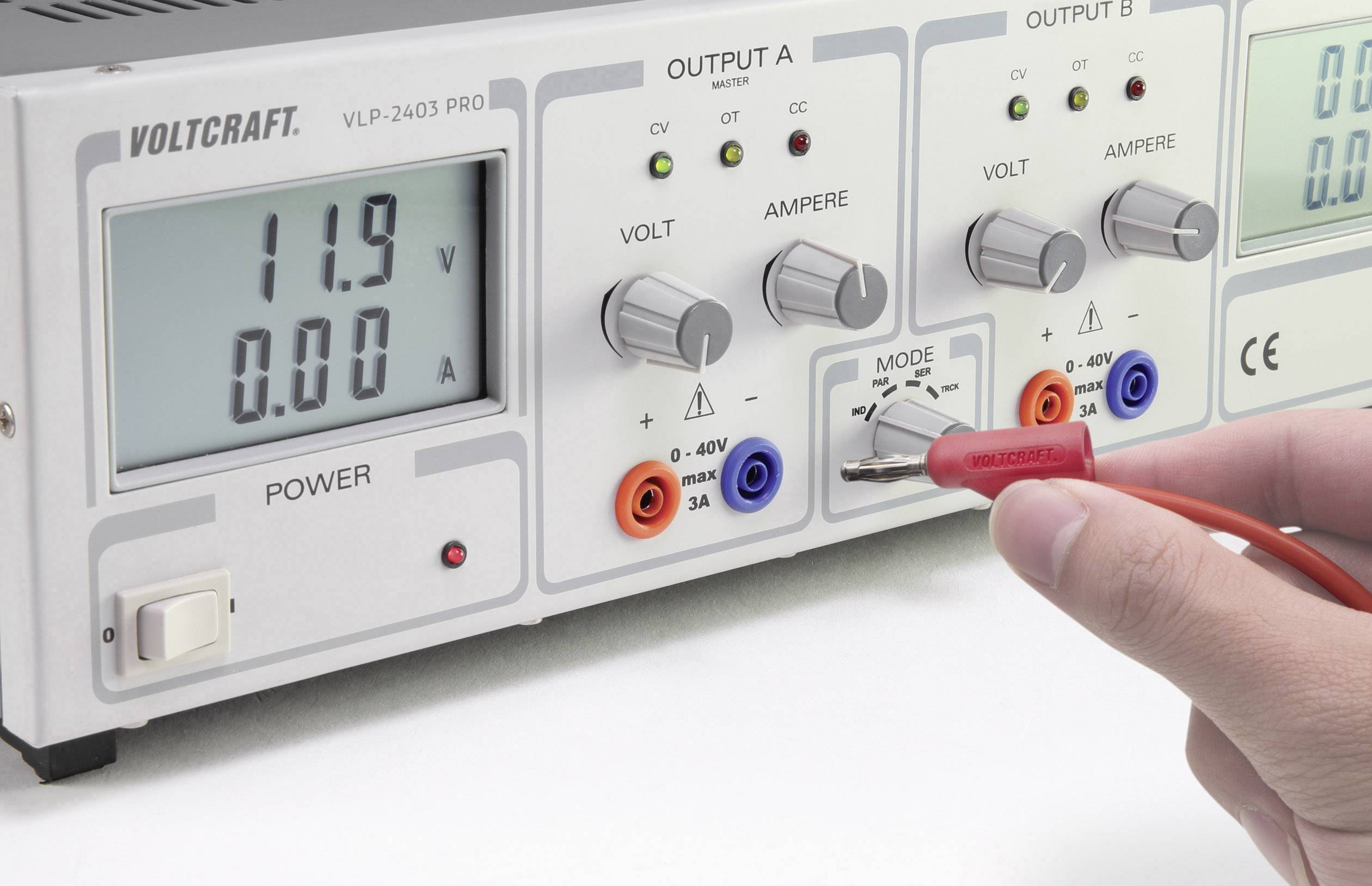Ruka drží červenou měřicí sondu u laboratorního síťového zdroje. Display ukazuje 11,9 V, 0,00 A. Jsou viditelné regulátory napětí a proudu a různé připojovací svorky.