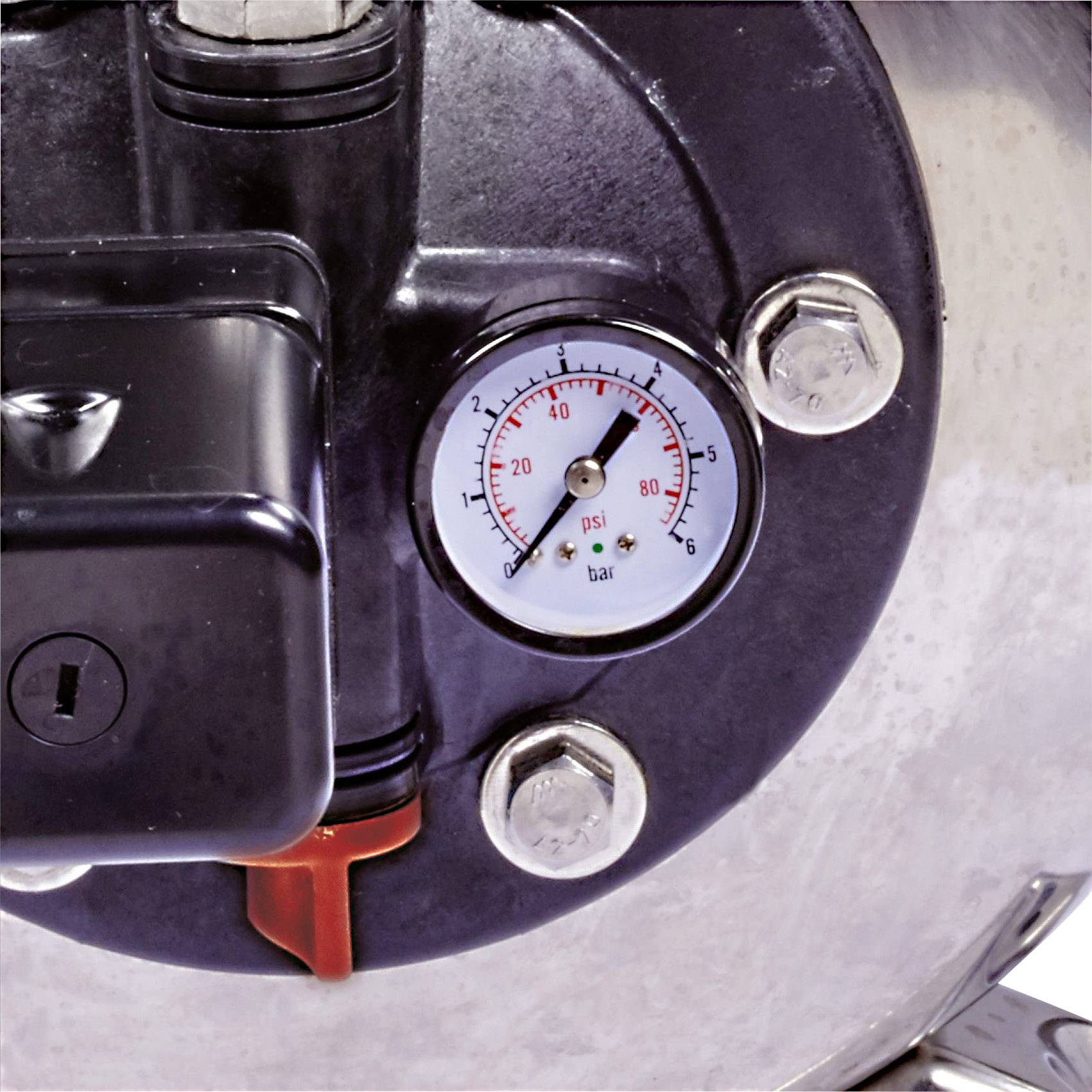 Manometr na kovovém kotli ukazuje tlak přibližně 20 psi. Manometr je obklopen několika šrouby.