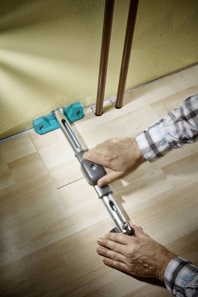 Osoba měří vzdálenost mezi stěnou a místem na dřevěné podlaze kovovým měřicím nástrojem.