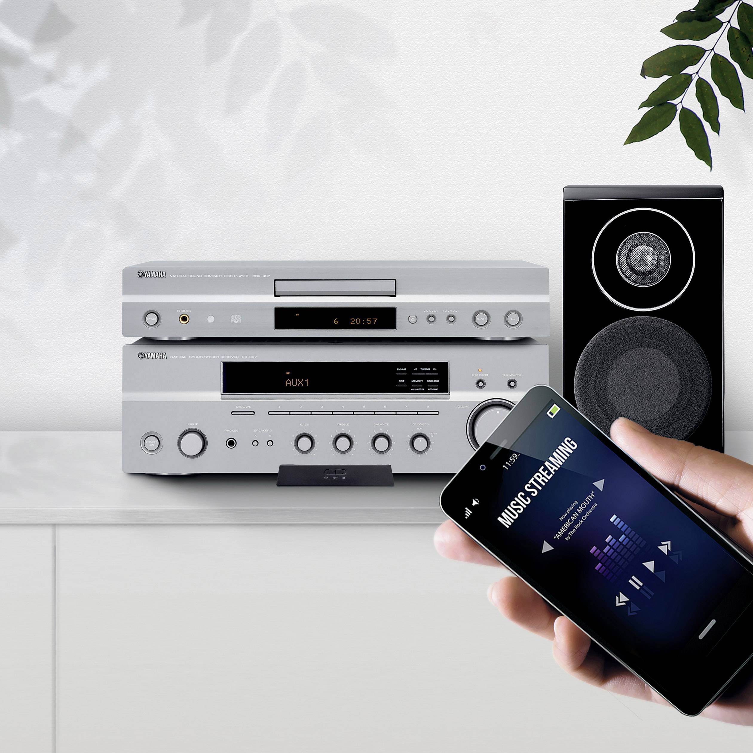 V popředí drží ruka smartphone zobrazující hudební streaming. V pozadí je sestava stereo systému s reproduktory.