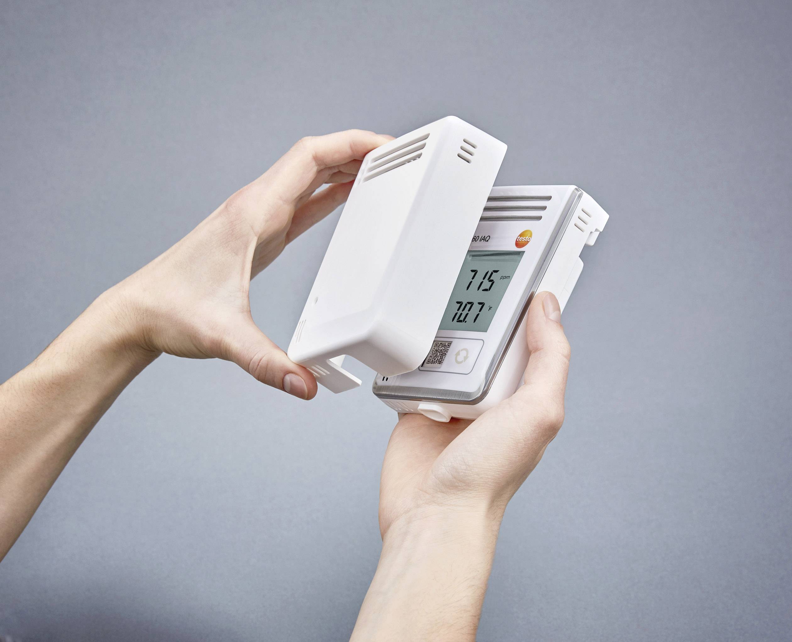 Osoba drží v rukou termostat. Display ukazuje teplotu '21,5°C'. Zařízení je v bílém pouzdře.