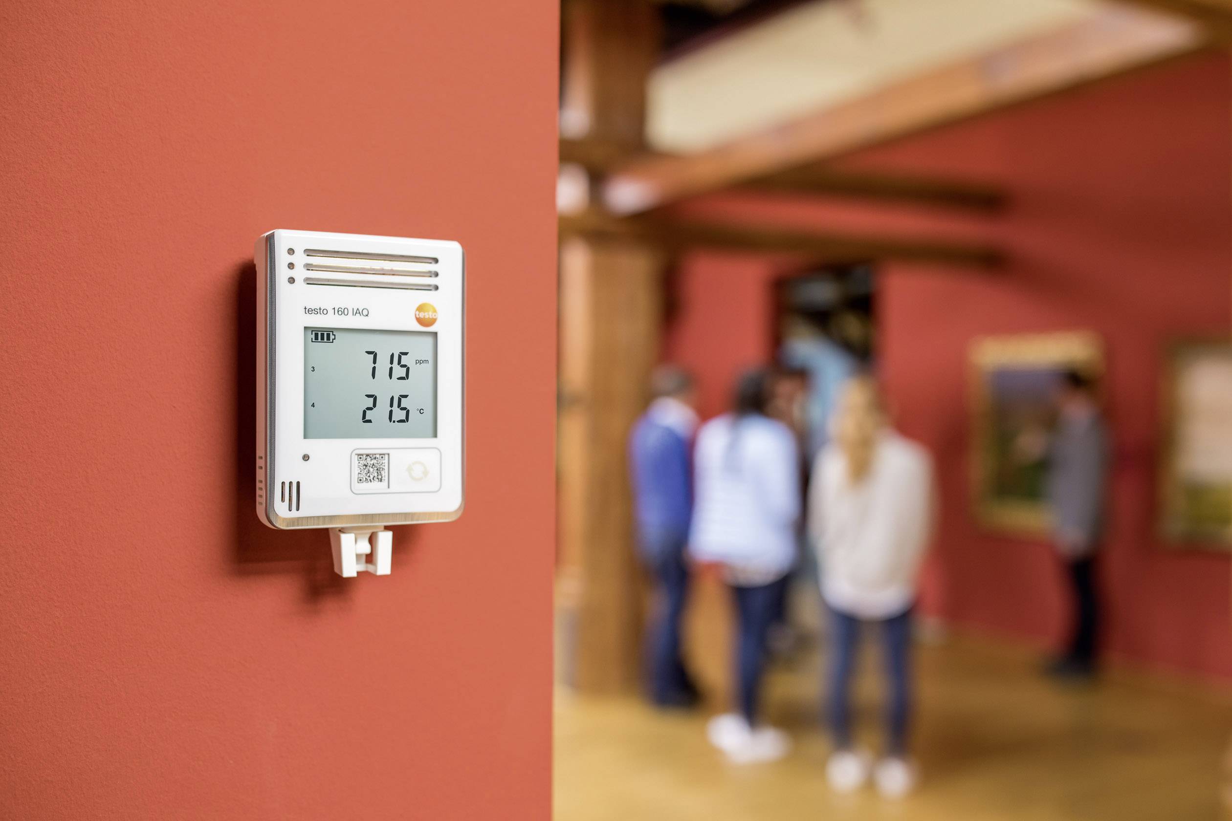 Digitální teploměr a vlhkoměr umístěný na červené stěně zobrazuje '21,5°' a '71,5%'. V pozadí jsou lidé v místnosti.