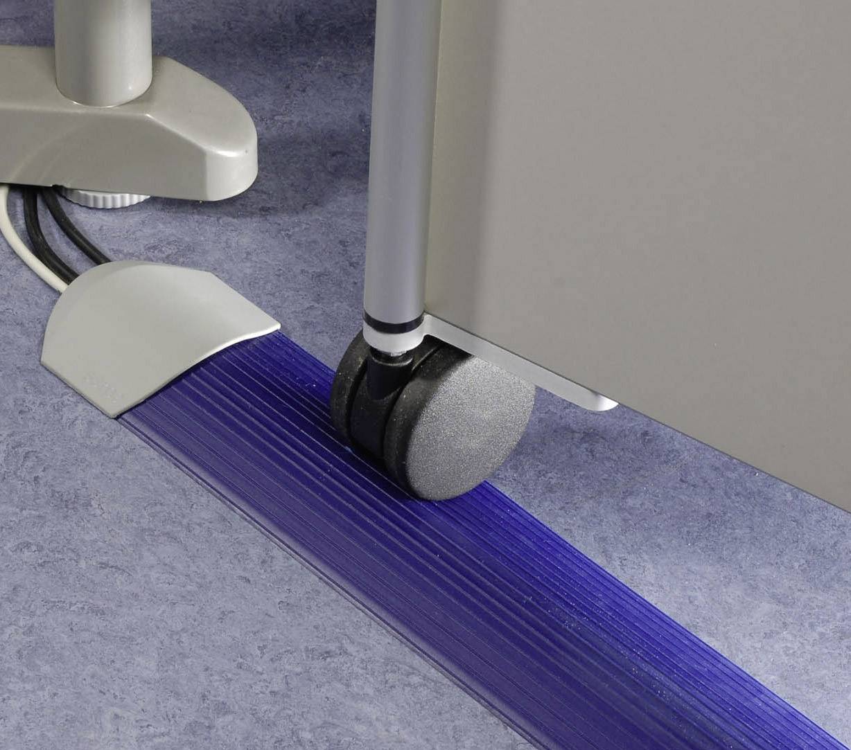 Kolečko kancelářské židle na modrém kabelovém kanálu, který uspořádává a chrání kabely na podlaze. Pozadí: šedá podlahová krytina a nábytek.