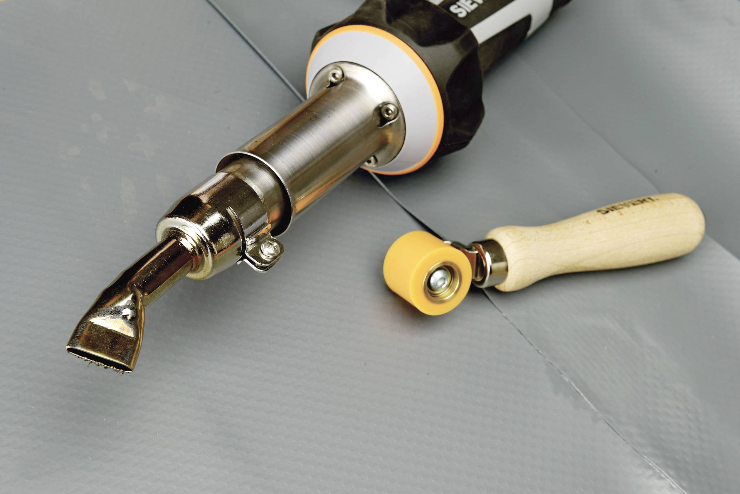 Horkovzdušný ventilátor se širokou tryskou vedle ruční svářečky na šedé plachtě, používaný pro svařovací práce ve zpracování plastů.