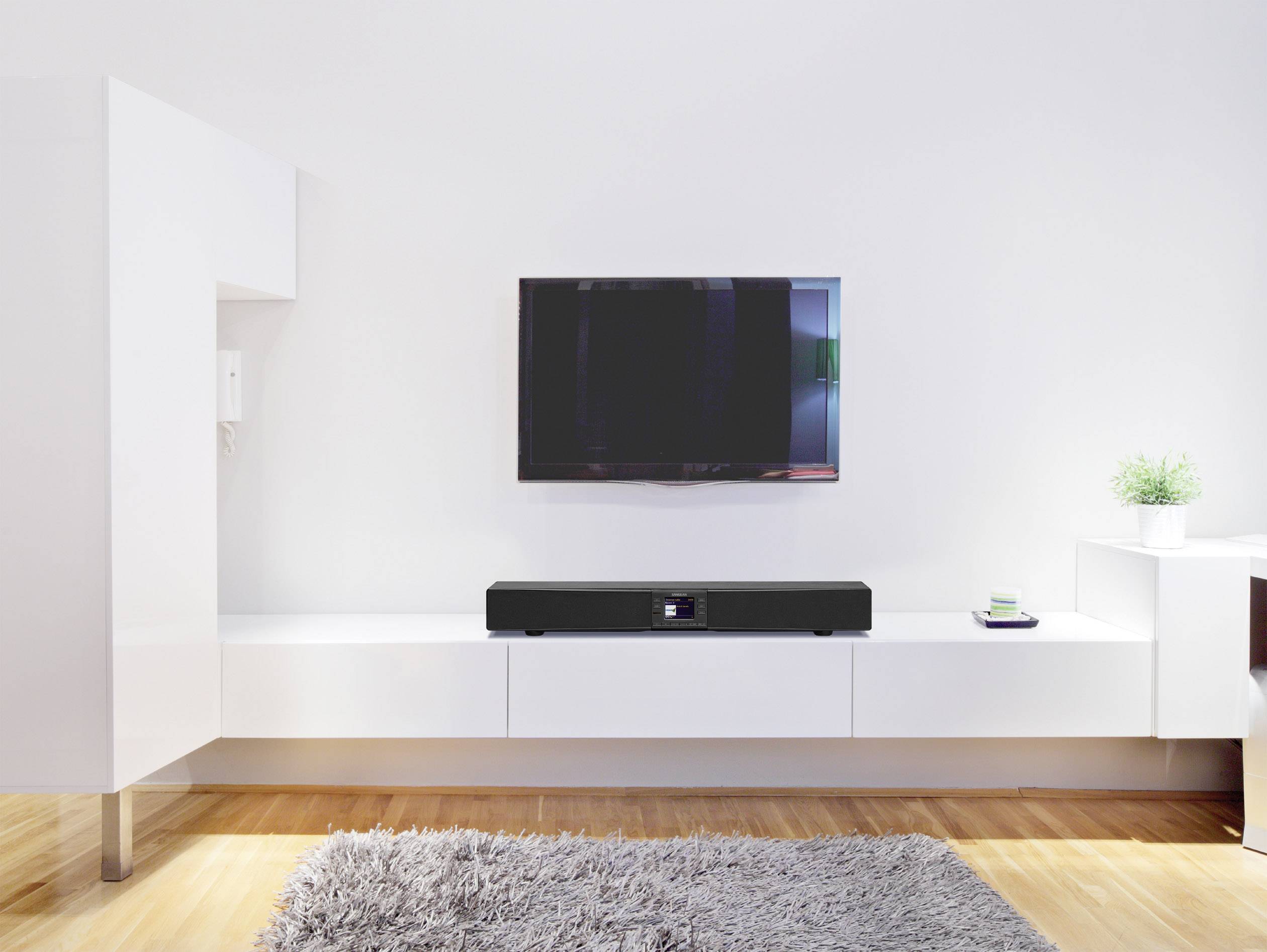 Obývací pokoj s moderní TV na stěně; pod ní je umístěna soundbar na bílém nízkém stolku. Koberec na dřevěné podlaze, minimalistické zařízení.