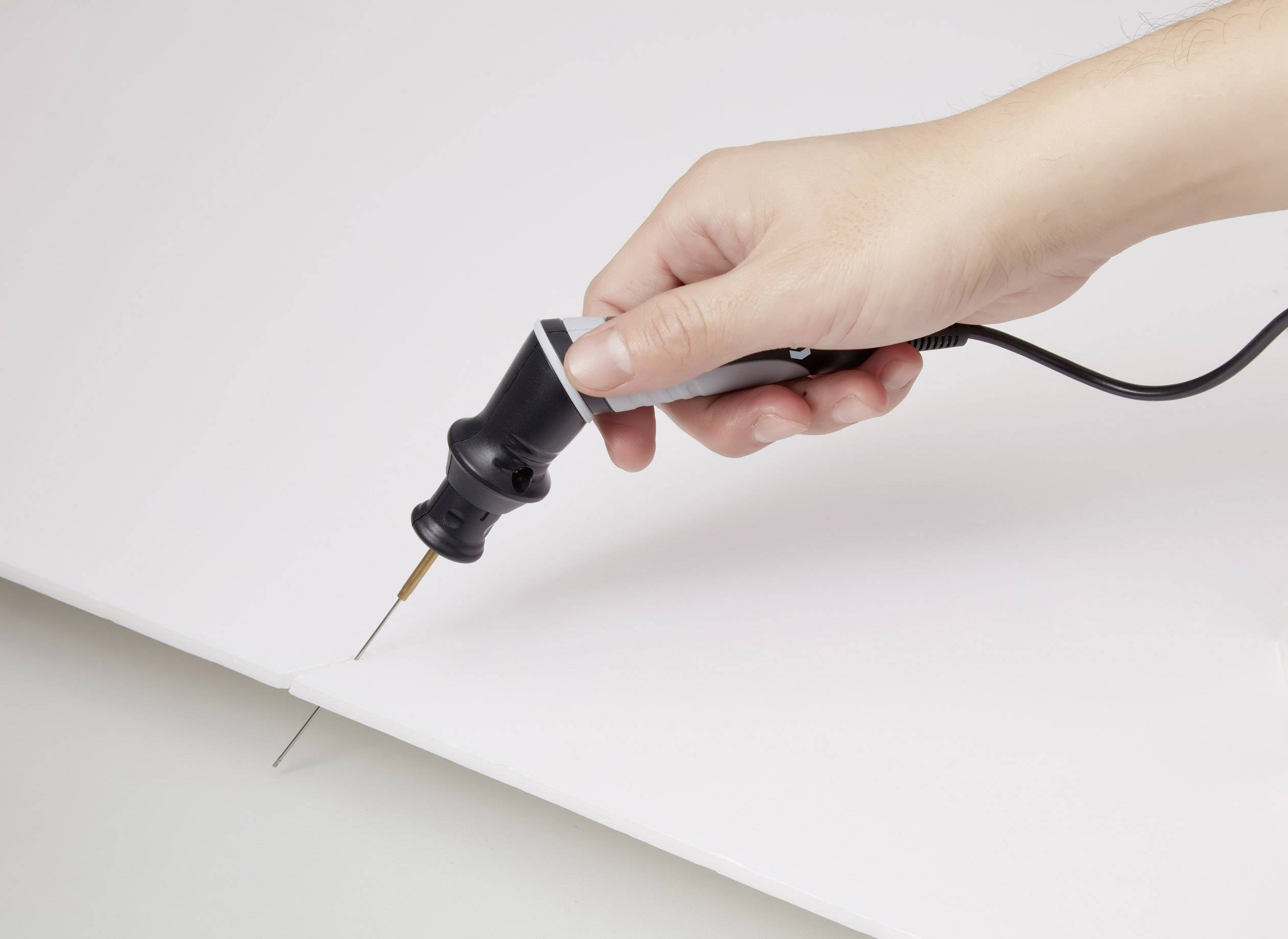 Jedna ruka drží elektrický nástroj s jemným hrotem, který řeže do bílého materiálu.