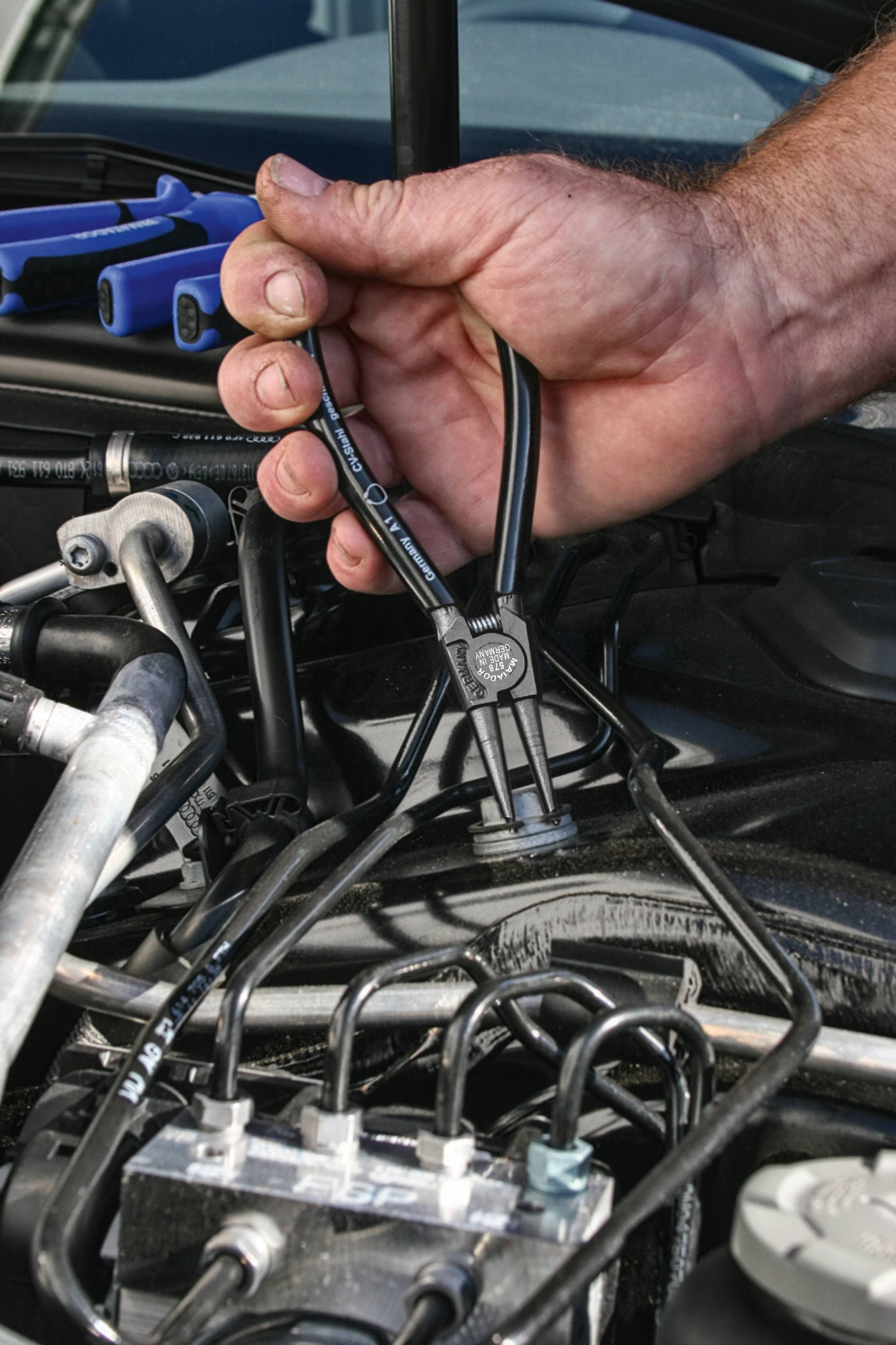 Jedna ruka drží stahovák, aby uvolnila svorku v motorovém prostoru automobilu. Mechanické nástroje a kabely jsou viditelné.