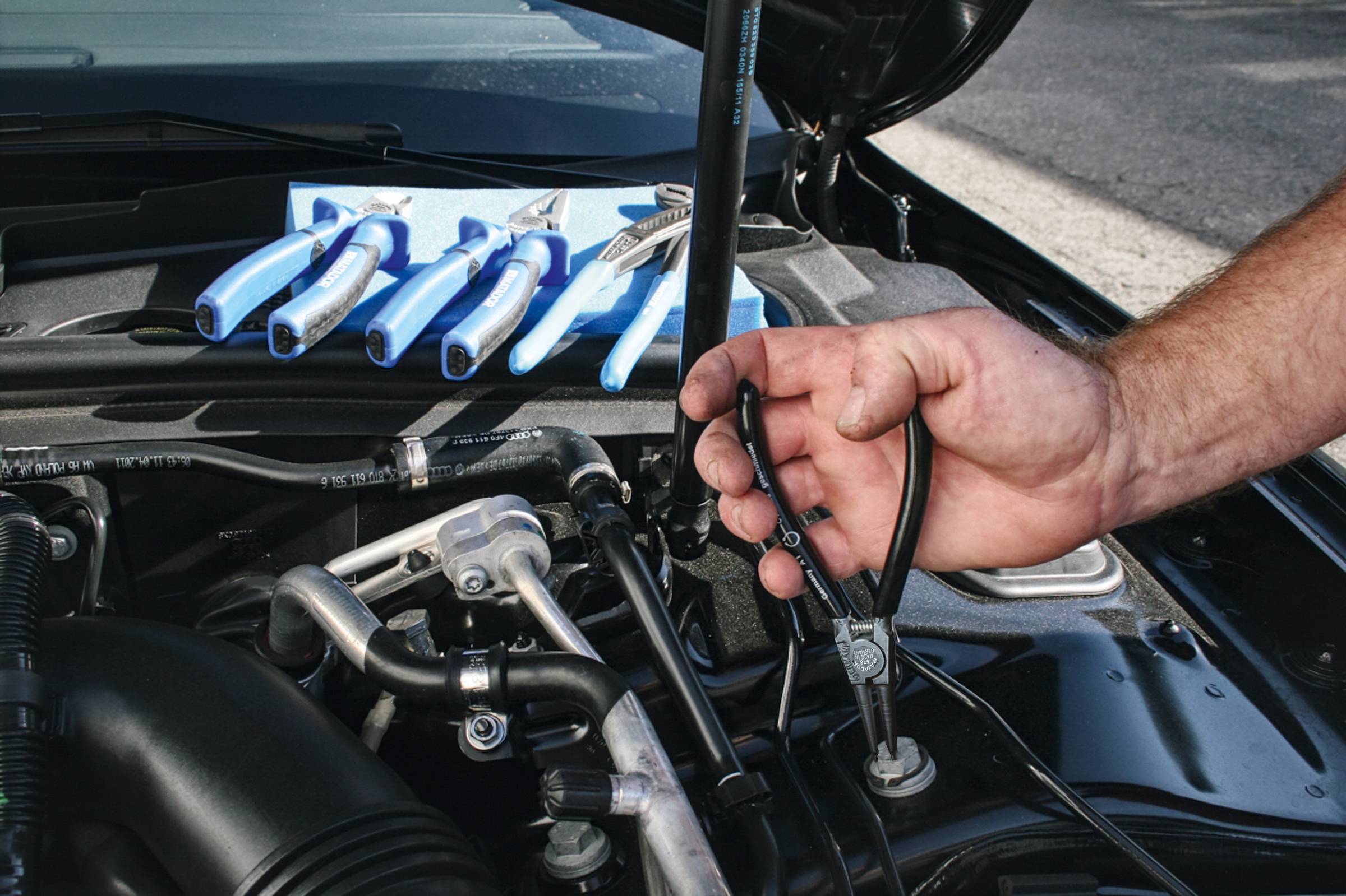 Mechanik drží kleště v motorovém prostoru automobilu. V pozadí leží řada nástrojů (šroubováků) na modrém plátně.