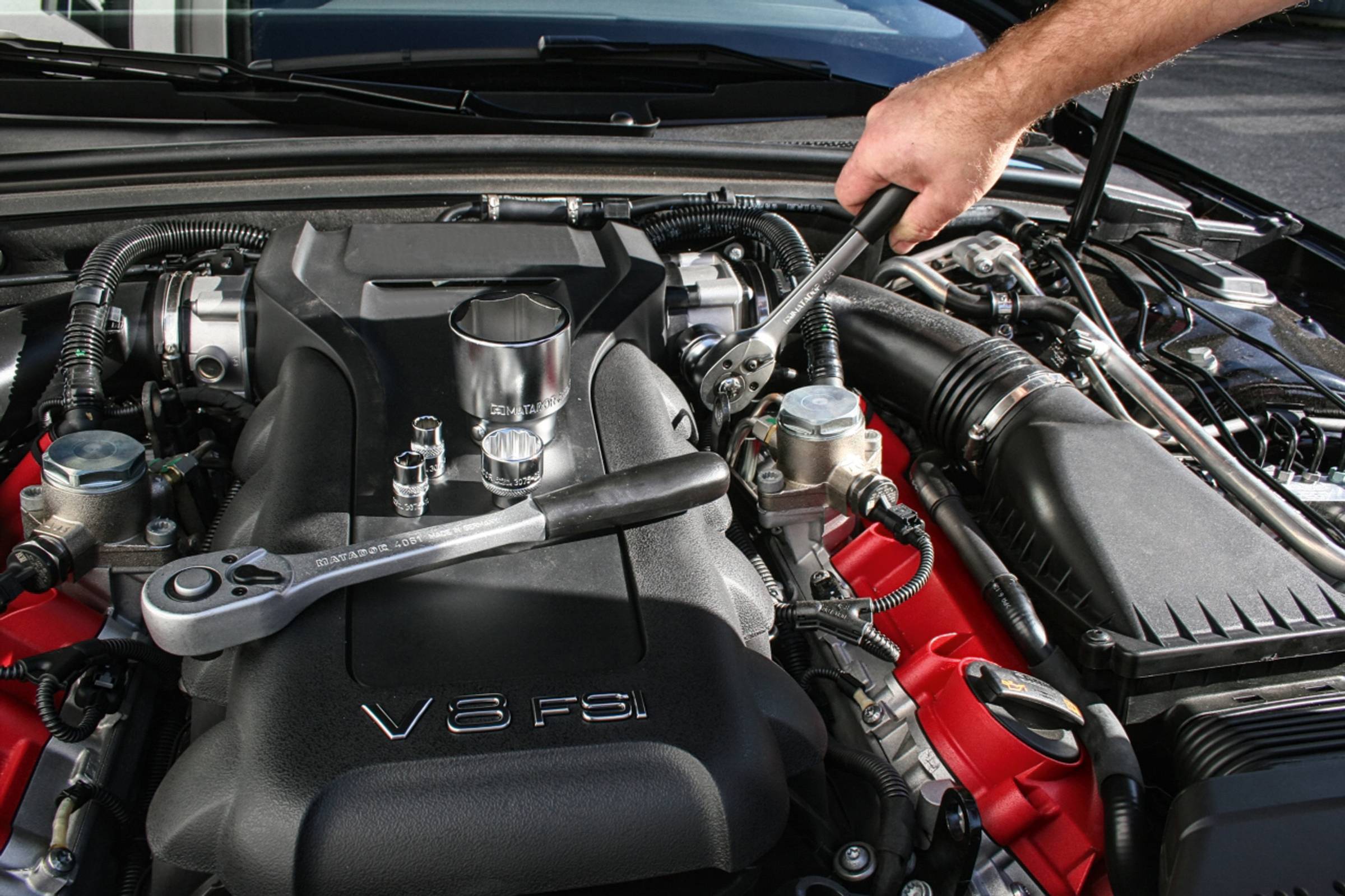 Ruka opravuje osmiválcový motor pomocí klíče. Motorový prostor je čistý a nářadí je připraveno.