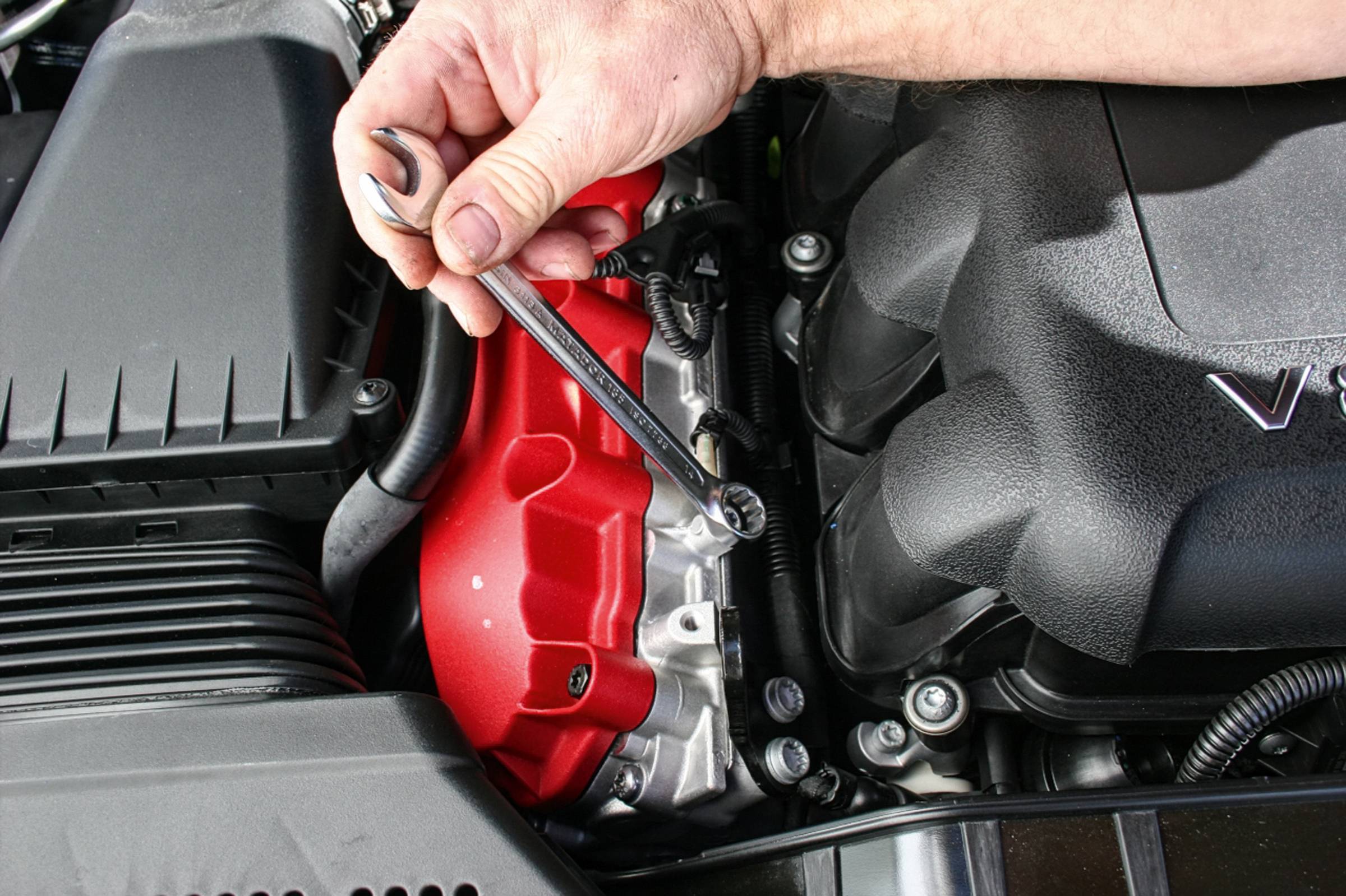 Osoba provádí údržbu červeného motorového krytu vozidla pomocí klíče, obklopena různými díly motoru.