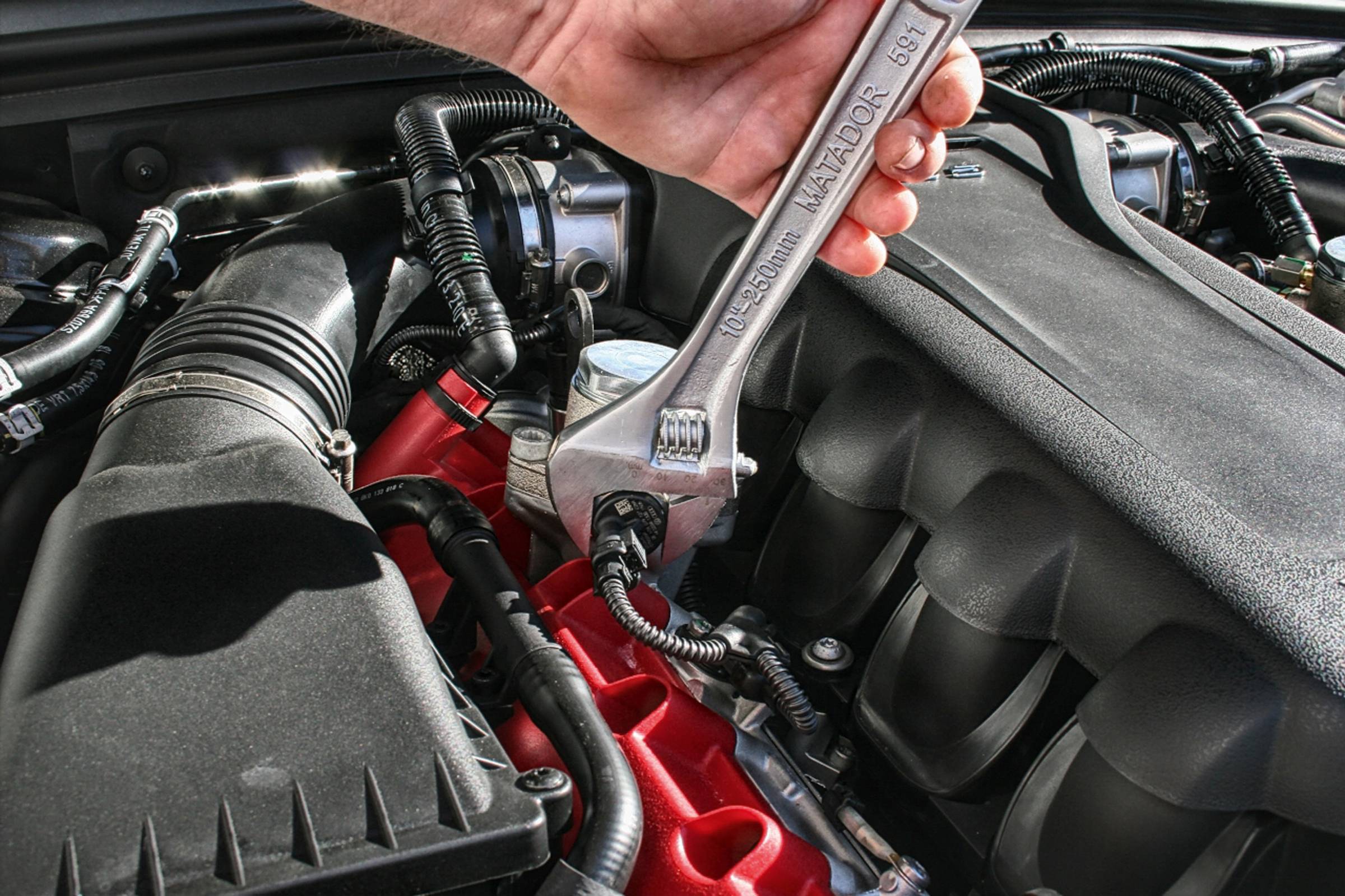 Ruka drží klíč, aby utáhla šroub v motorovém prostoru automobilu. Motorový prostor ukazuje různé kabely a komponenty.