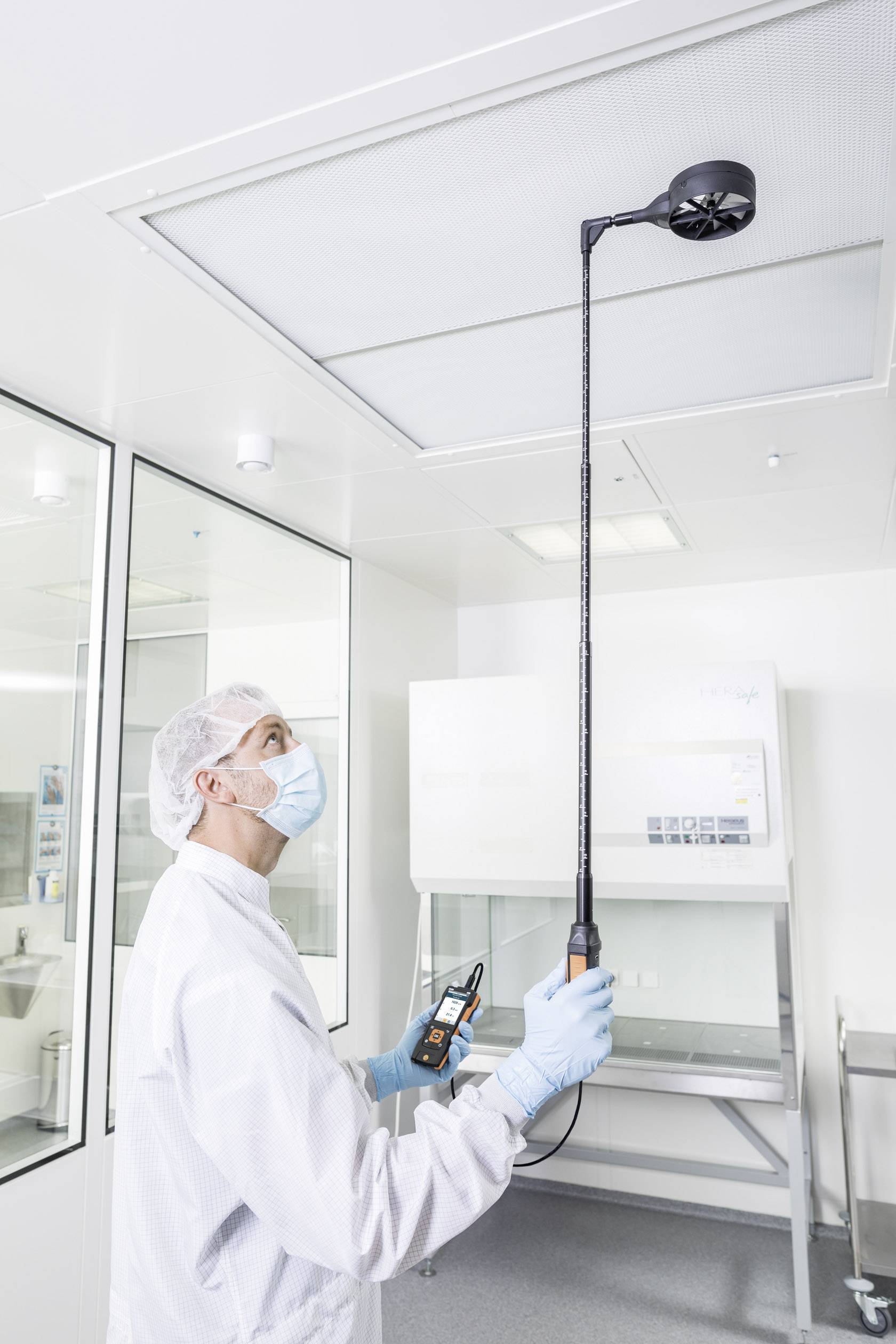 Osoba v ochranném oblečení měří proudění vzduchu v laboratoři pomocí ruční měřicí jednotky a sondy umístěné na stropě.