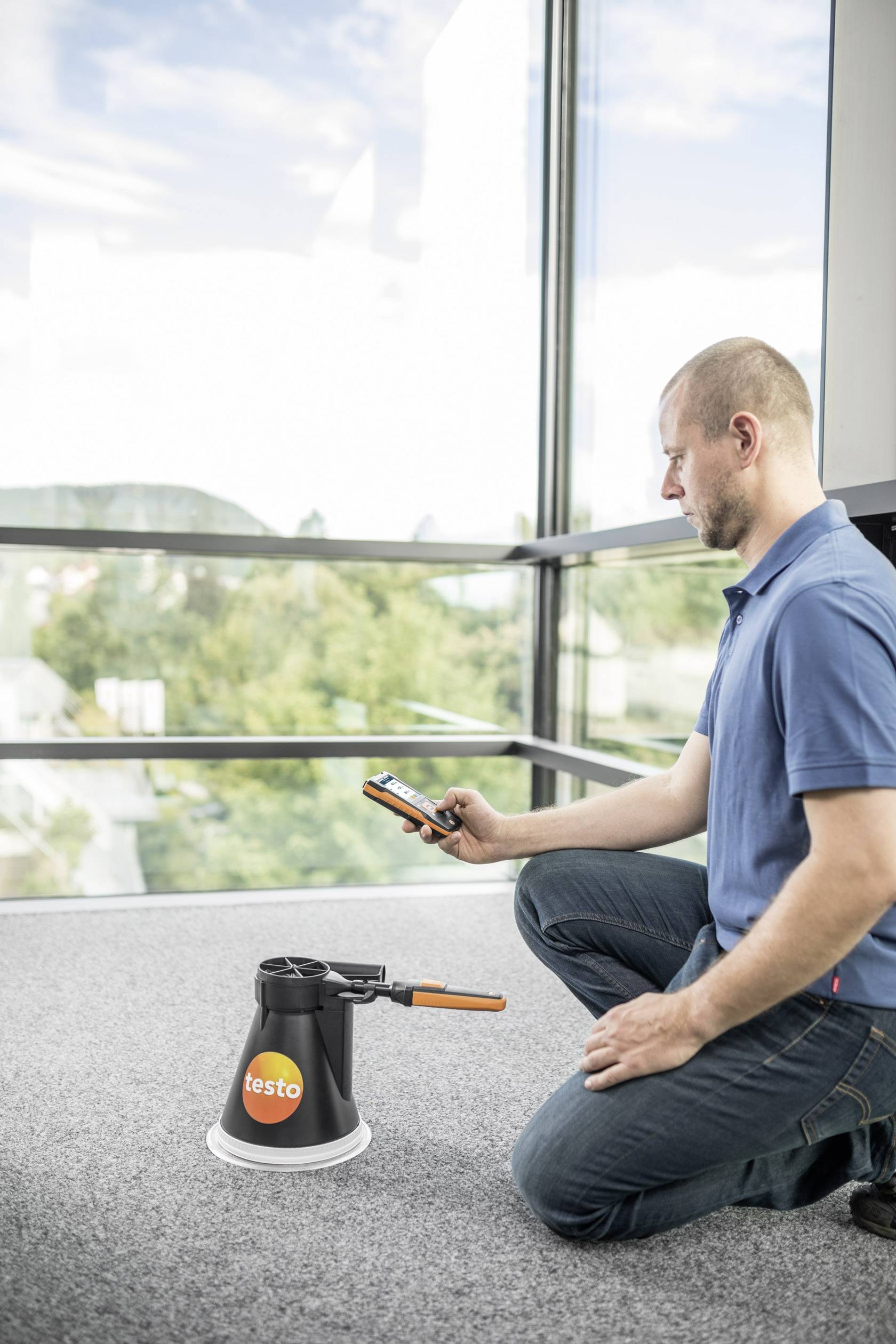 Muž klečí na podlaze a drží měřicí přístroj. Před ním stojí zařízení značky 'testo' pro měření kvality vzduchu v místnosti.