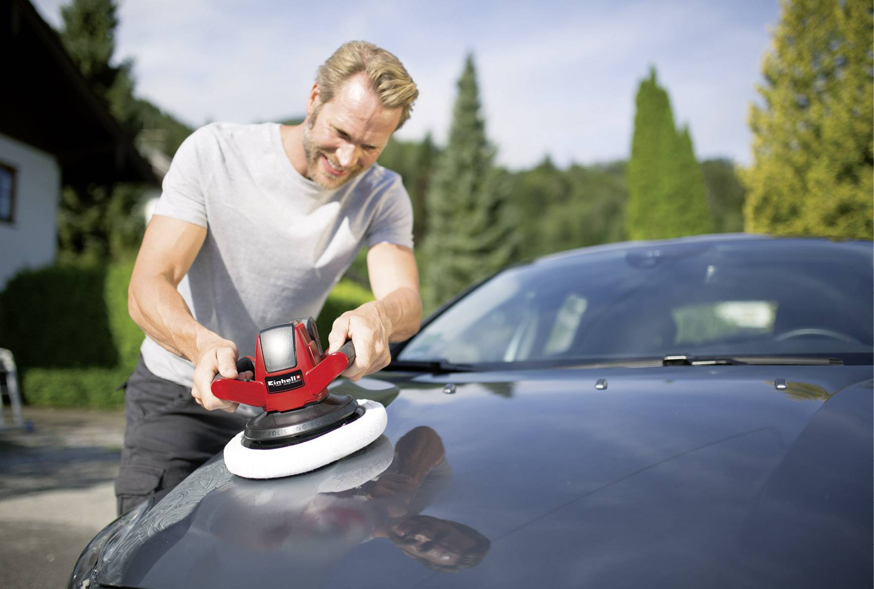 Muž leští kapotu automobilu leštičkou venku. V pozadí jsou vidět stromy a budova.