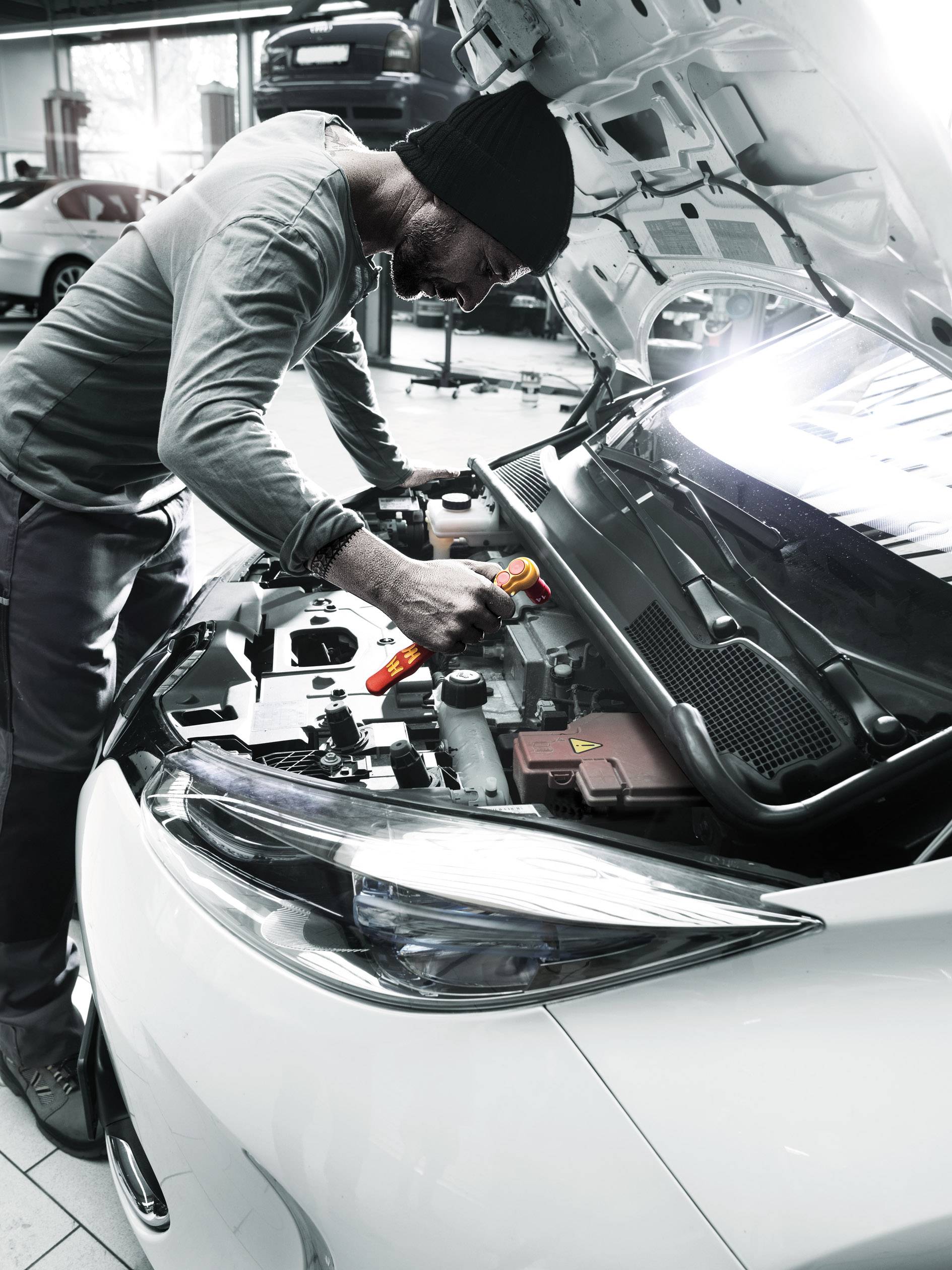 Muž opravuje motor v autoservisu. Má na hlavě čepici a drží nástroj. V pozadí je další automobil.