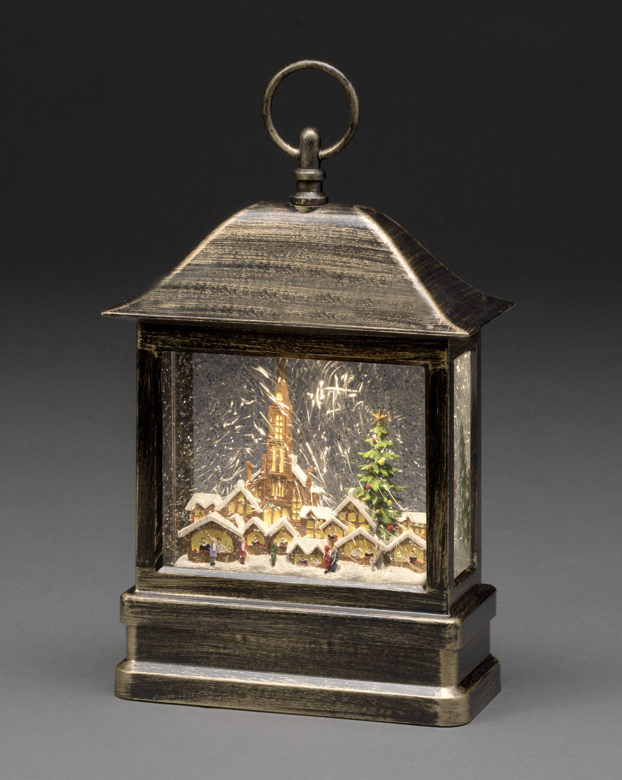 Lucerna zobrazuje zimní scénu s kostelem, vánočním stromkem a sněhem pokrytými domy, obklopené padajícím sněhem.