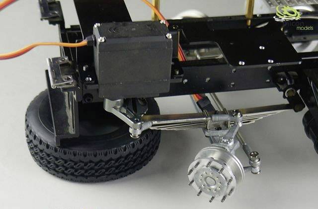 Detailní záběr modelové nápravy nákladního vozidla s pneumatikou a servopohonem, zobrazující mechanické detaily zavěšení kola.