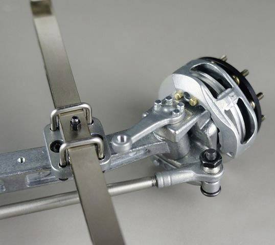 Kovový mechanický přístroj s pákou a řemenem, pravděpodobně součást nějakého zařízení nebo stroje, na šedém pozadí.