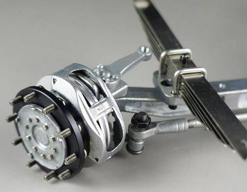Detailní záběr brzdového systému vozidla s kotoučovou brzdou a listovou pružinou, odhalující mechanické detaily a propojení.