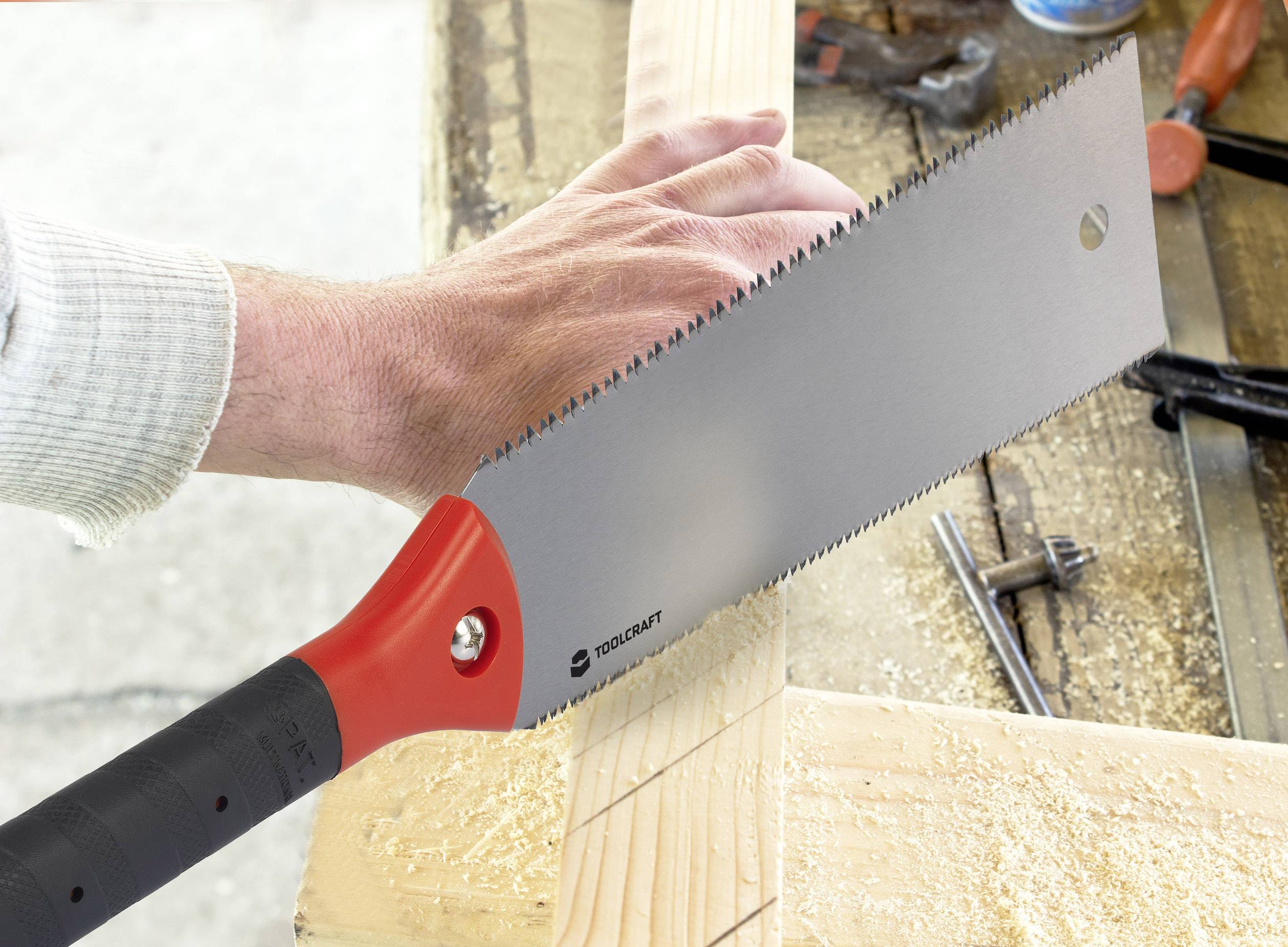 Osoba řeže dřevěné prkno ruční pilou podél vyznačené čáry v dílně. Piliny leží na pracovním stole.
