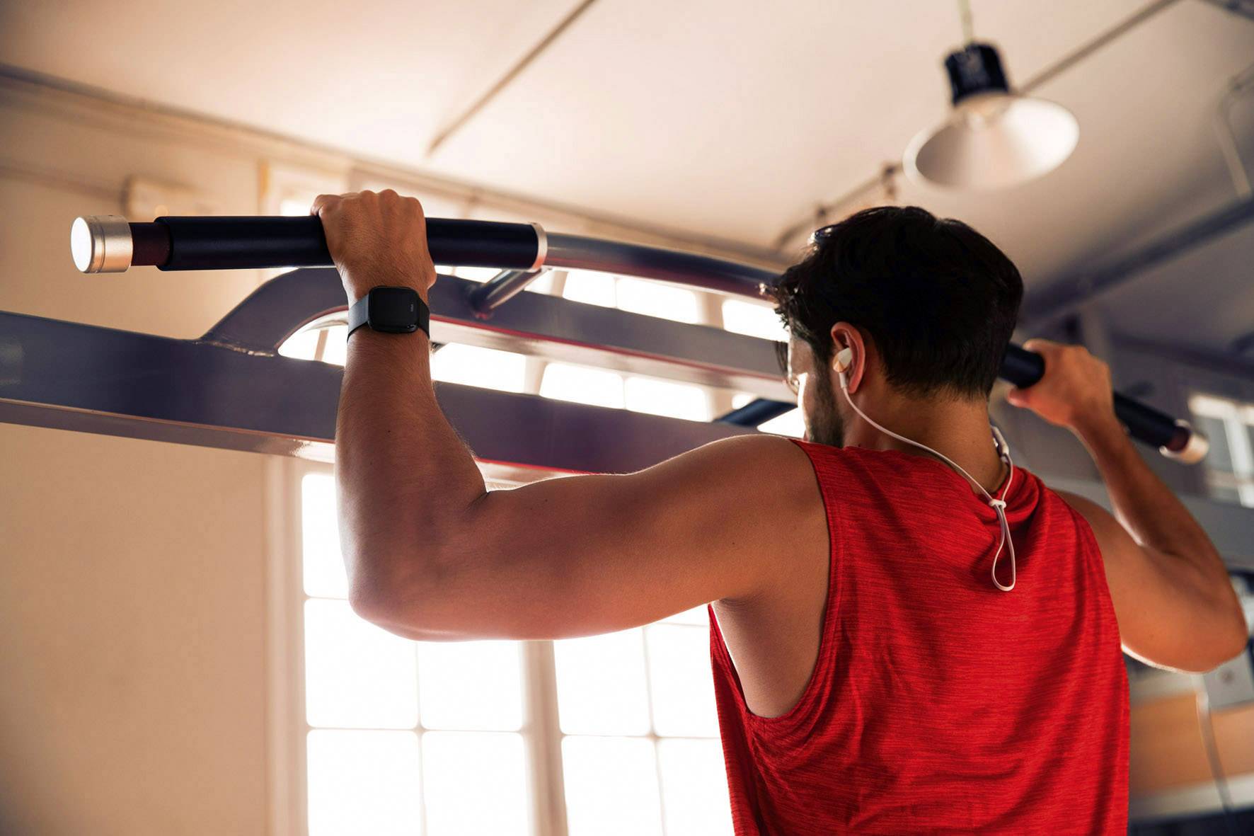 Muž v červeném tričku provádí shyby na tyči ve fitness centru. Má na uších sluchátka a na zápěstí fitness náramek.