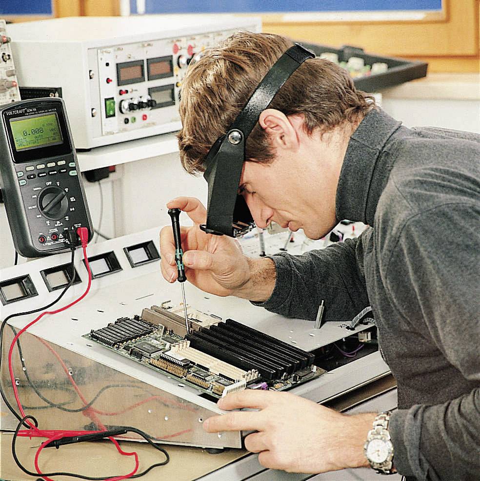 Technik pracuje šroubovákem na desce plošného spoje; poblíž multimetr měří elektrický odpor.