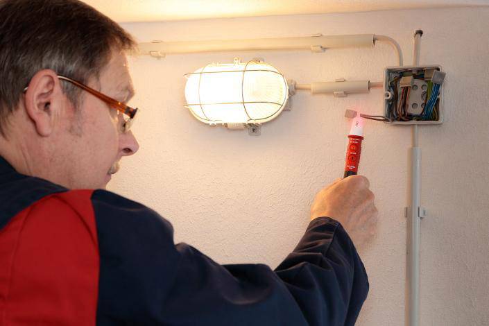 Elektrikář kontroluje zapojení vedle nástěnné lampy pomocí zkoušečky napětí, aby zajistil bezpečnost instalace.