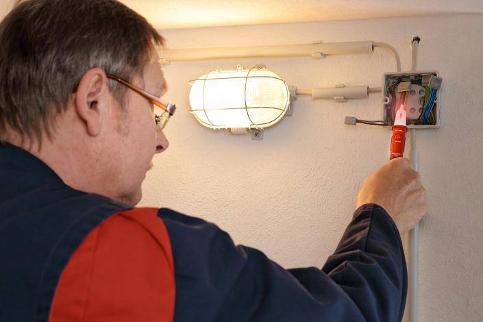 Elektrikář kontroluje otevřenou rozvodnou krabici pomocí zkoušečky napětí na zdi pod svítící lampou.