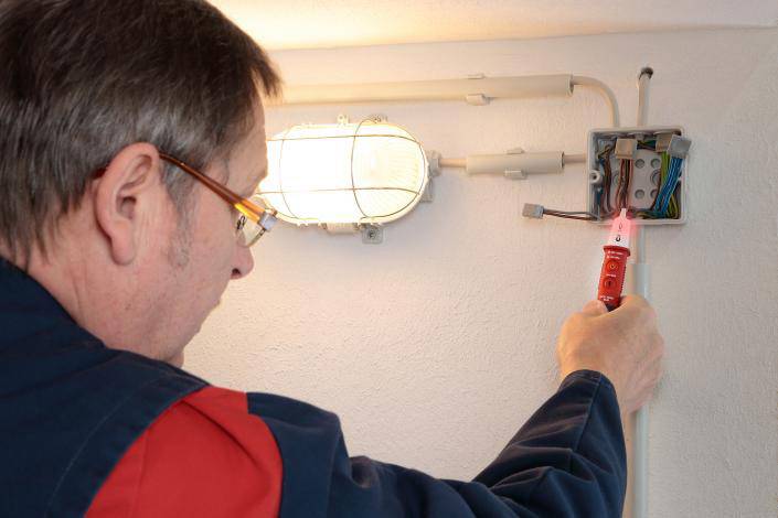 Elektrikář kontroluje zapojení v nástěnné zásuvce vedle lampy pomocí zkoušečky napětí.